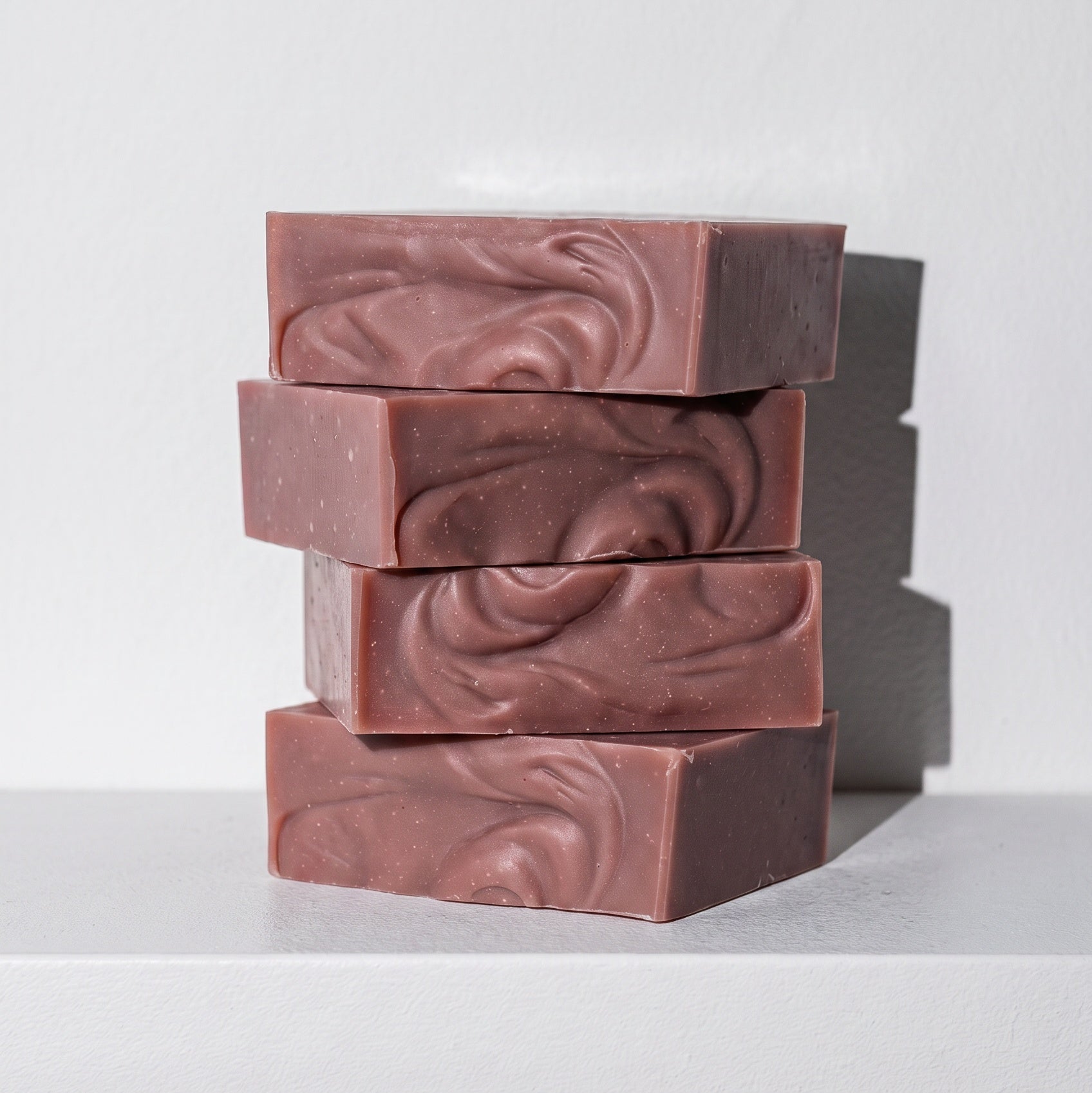 Four rectangular bars of handmade soap stacked vertically on a white shelf against a light gray wall. The soap has a dusty rose or mauve-brown color with a swirled, marbled texture on the top surface.