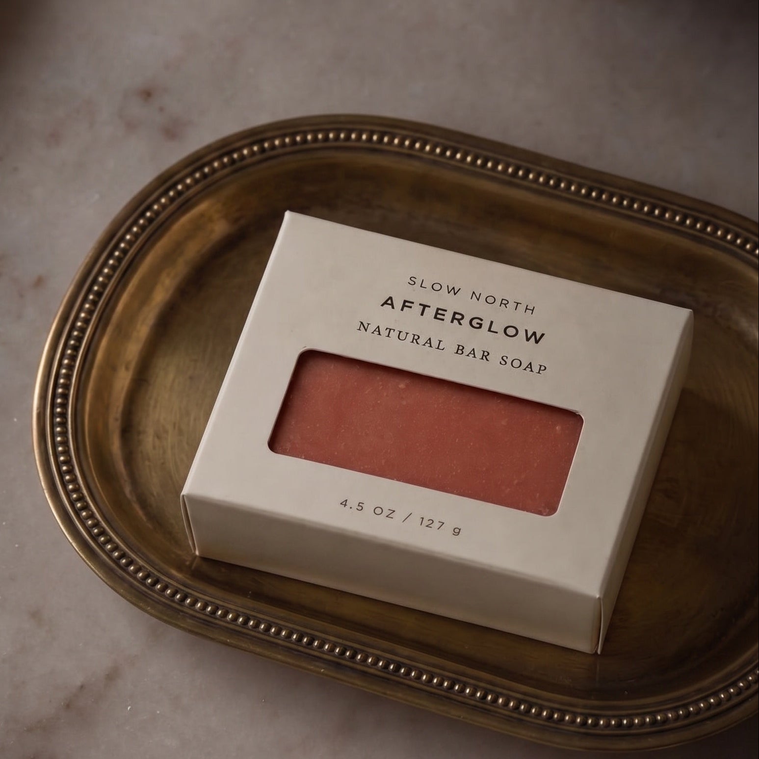 Slow North Afterglow natural bar soap in a cream-colored rectangular box with a clear window showing the coral-pink soap inside, resting on an oval gold tray.