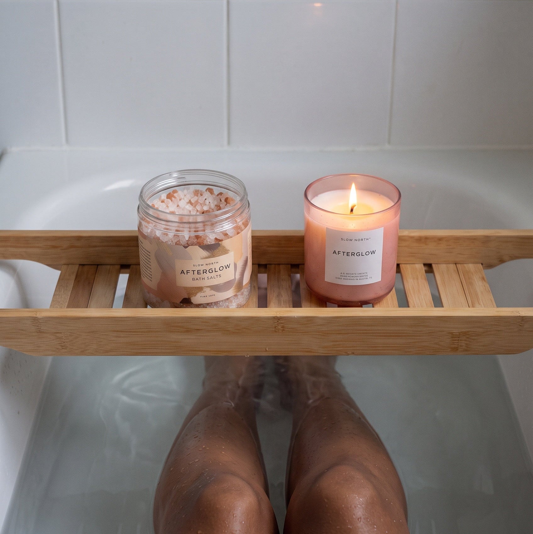 A bamboo bath caddy holding a pink bath salt jar and a lit pink candle, placed over a bathtub with a person's legs visible below the tray.