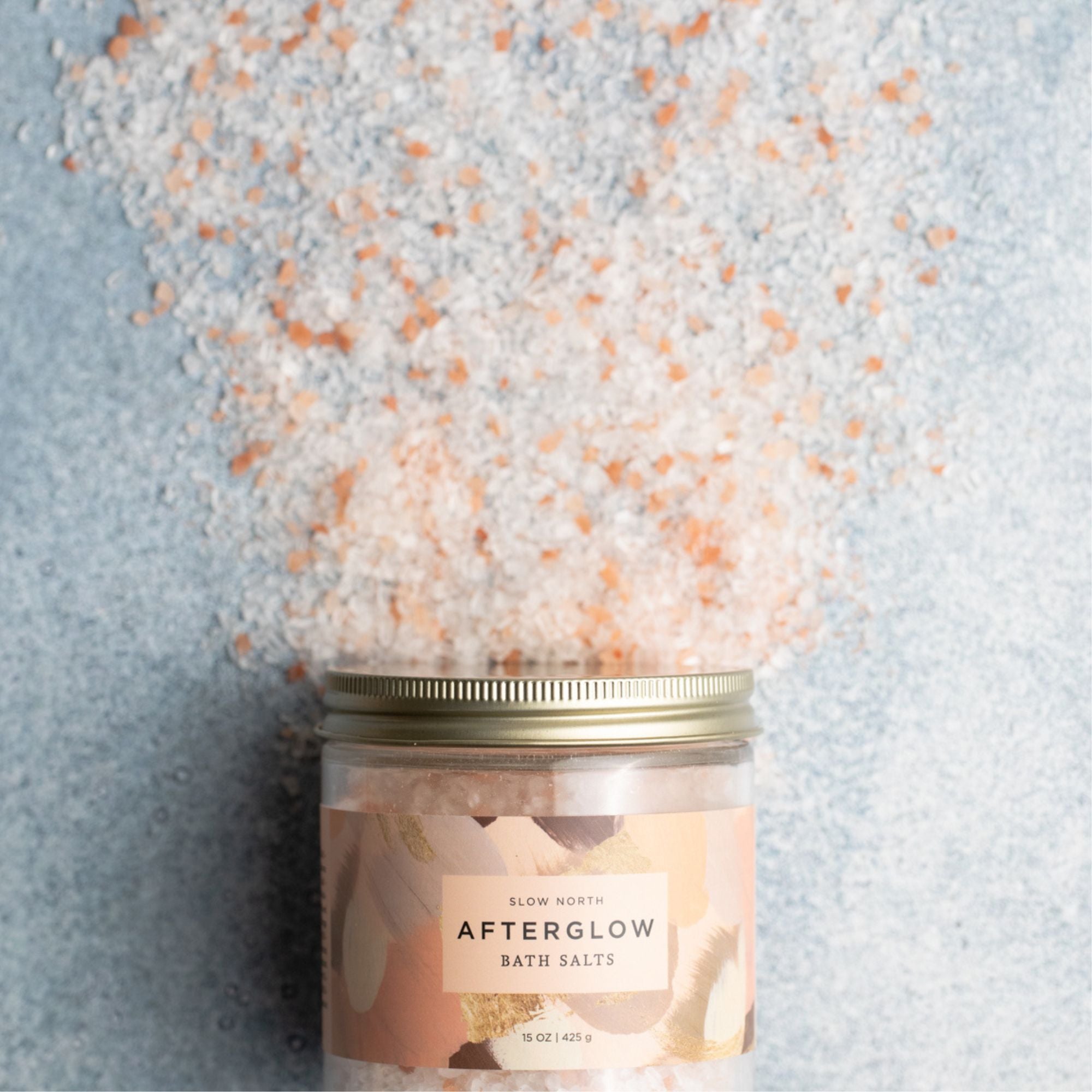 Jar of 'Afterglow' bath salts with pink and white bath salts spilling out on a light blue background
