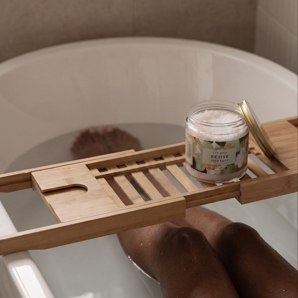 A wooden bath caddy draped over a white bathtub, holding a small jar of bath salts with a gold lid. A person's legs are visible in the water, and the background shows a tiled bathroom wall.