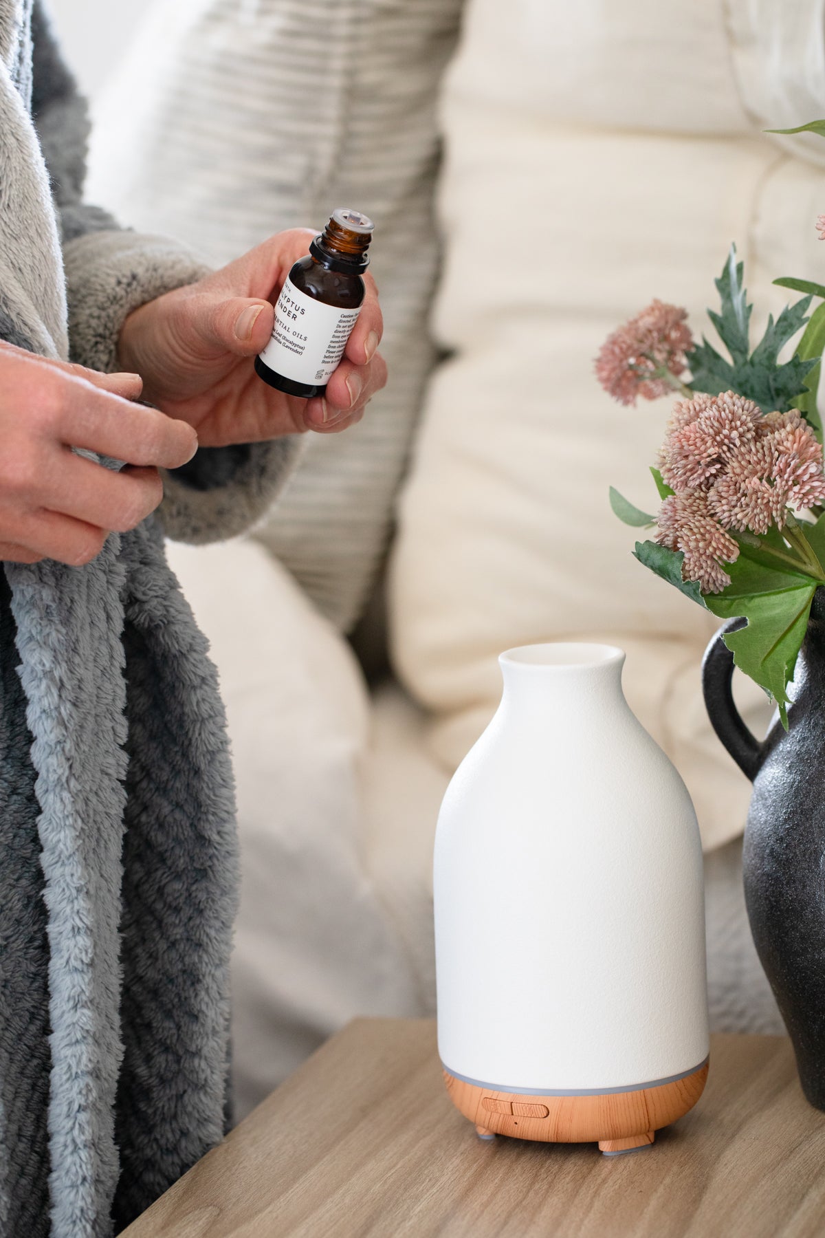 Person holding a bottle of essential oil next to a white diffuser on a wooden surface.