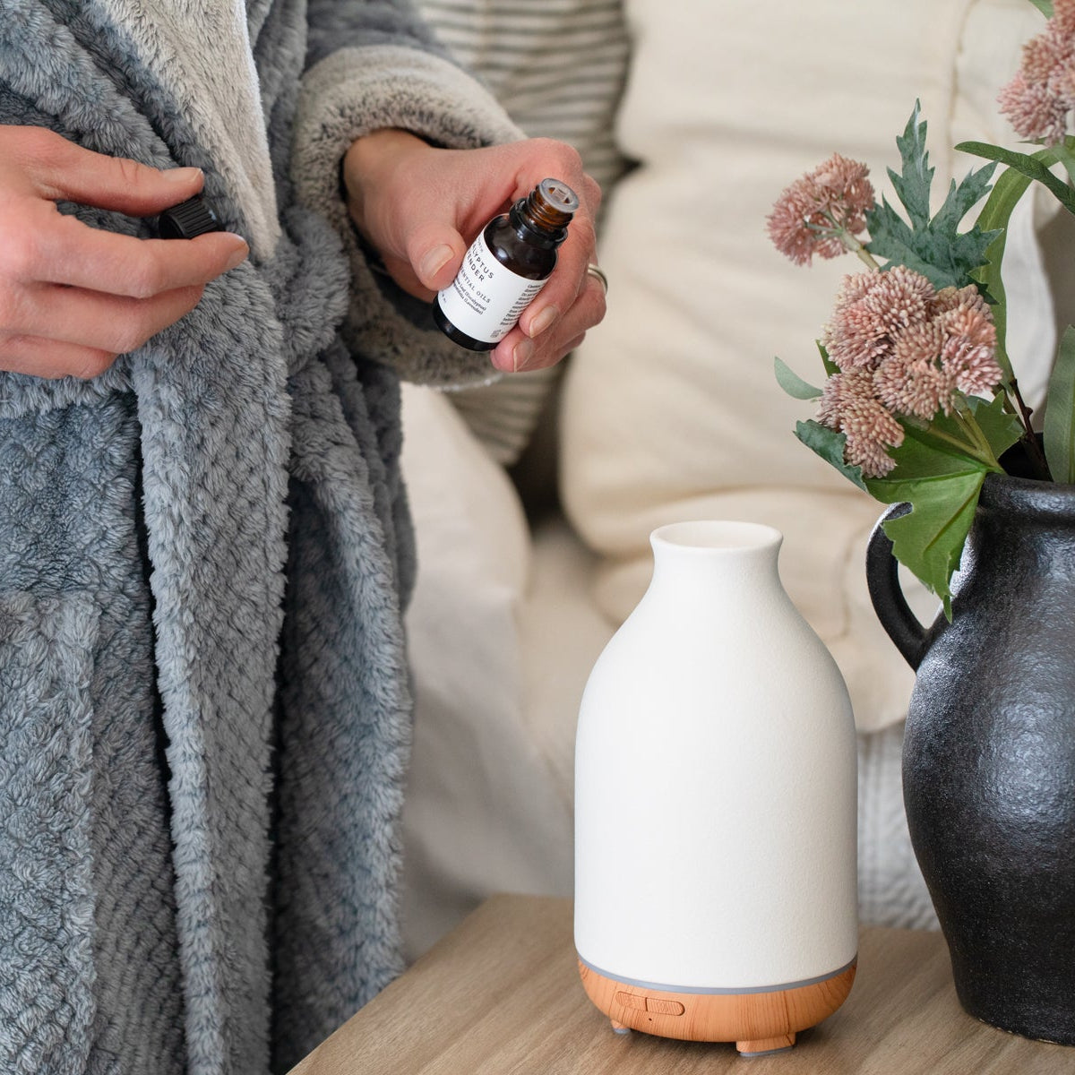 Person in a gray robe holding a bottle of essential oil next to a diffuser on a table.