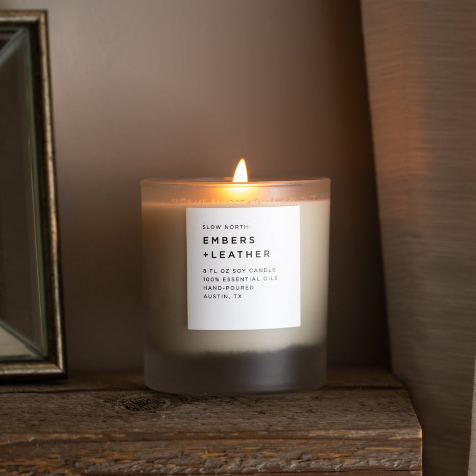 Candle labeled 'Ember & Leather' on a wooden surface with a curtain in the background