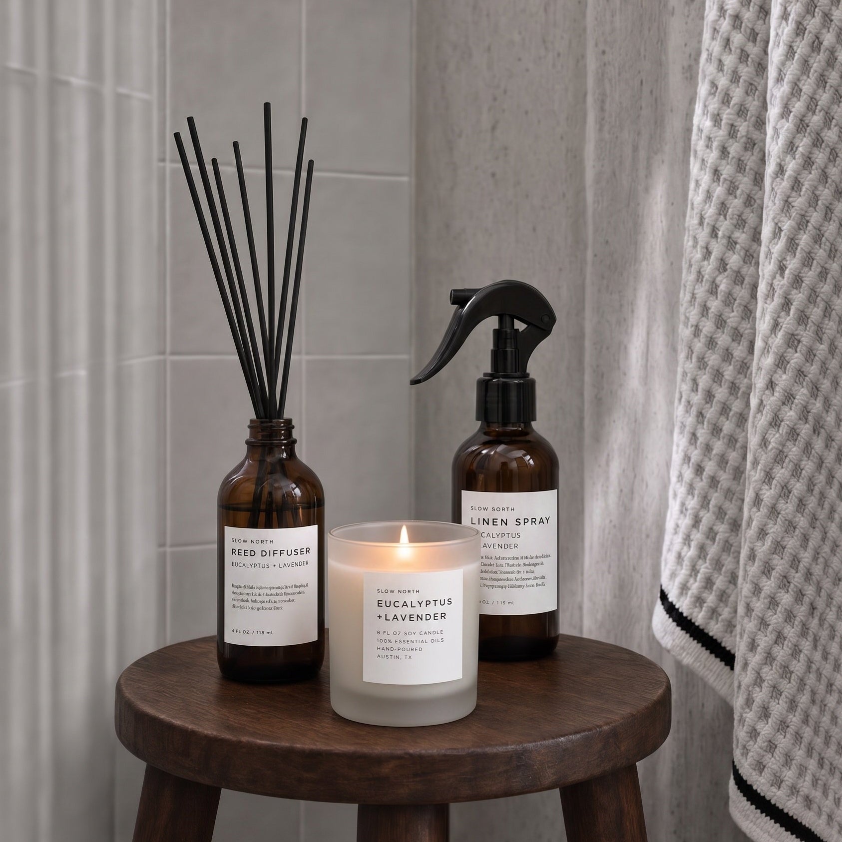 Three home fragrance products arranged on a round wooden stool in a bathroom setting: an amber glass reed diffuser bottle with black reeds, a lit white candle in a frosted glass holder, and an amber glass linen spray bottle with a black sprayer. A gray tiled wall and a patterned towel are visible in the background.