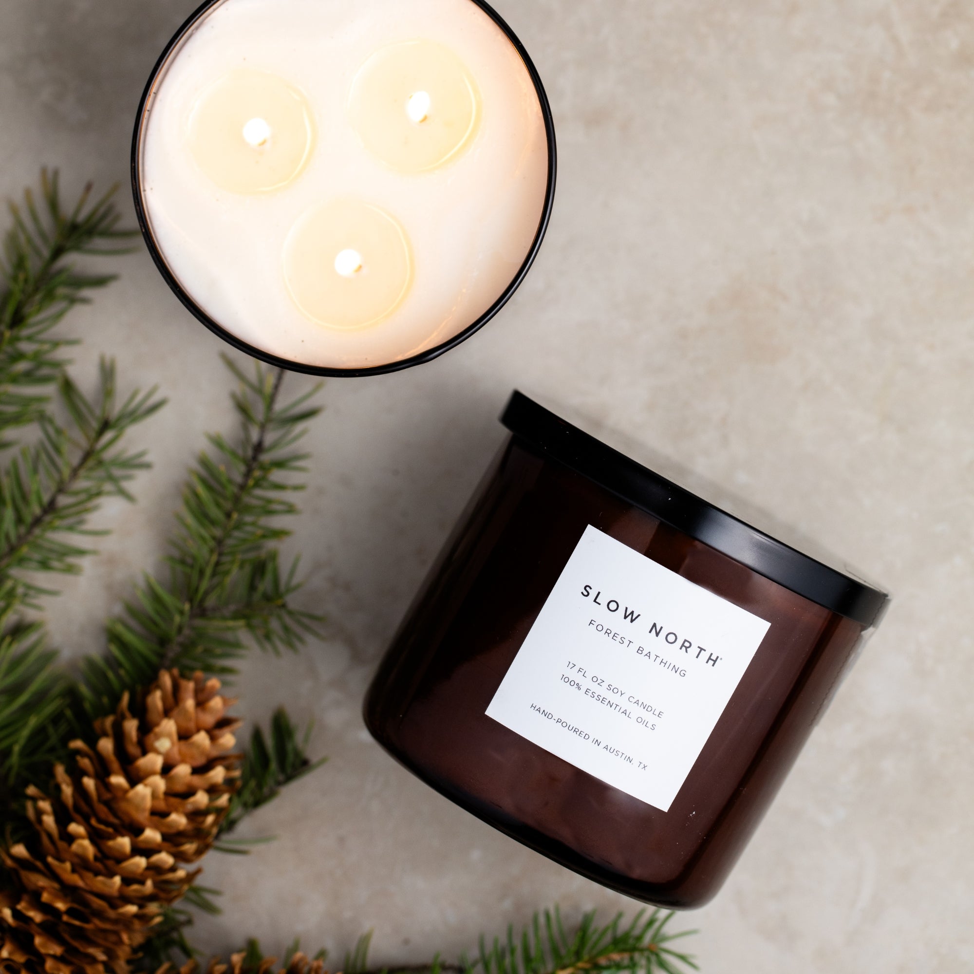 Candle with a visible brand label surrounded by pine branches and a pine cone on a textured surface.
