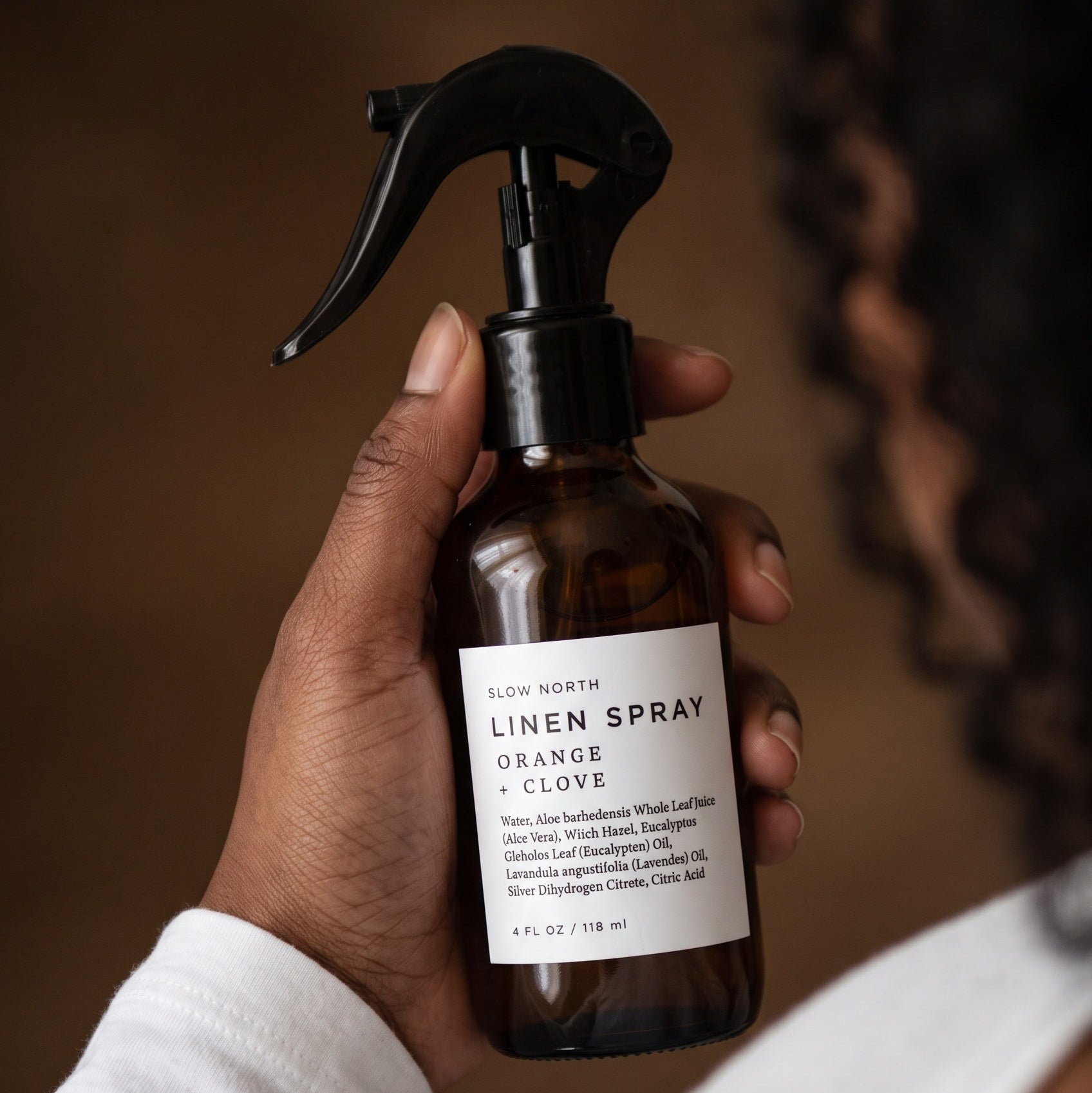 A hand holding an amber glass spray bottle of Slow North Linen Spray in Orange Clove, with a person wearing a white long-sleeve top in the blurred background.