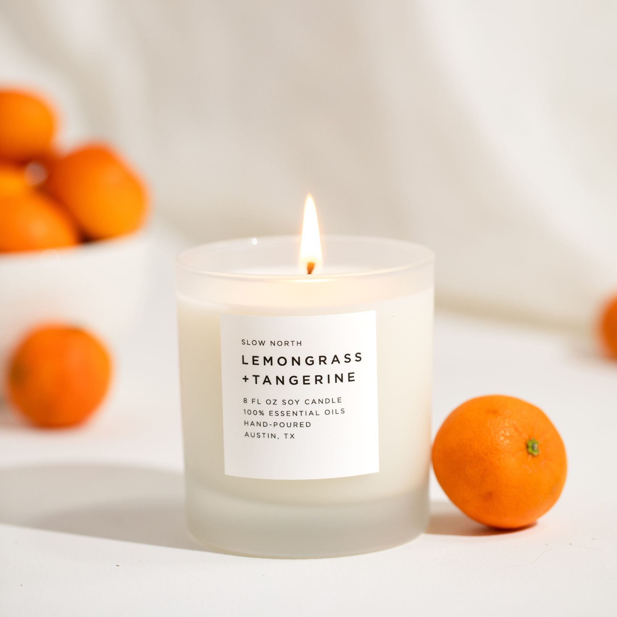 Candle labeled 'Lemongrass + Tangerine' with tangerines around on a light background