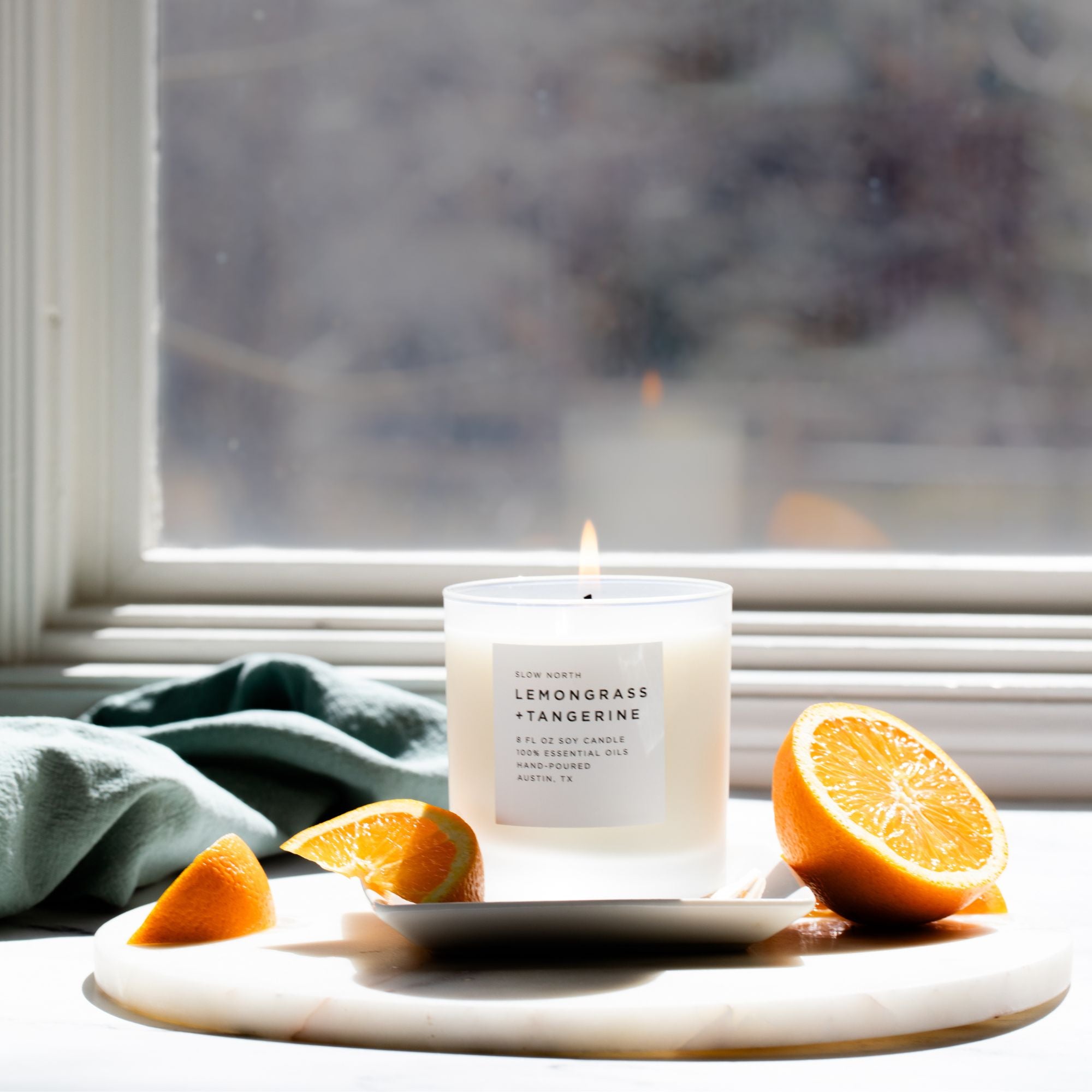Candle with lemongrass and tangerine scent on a marble tray with orange slices, near a window.