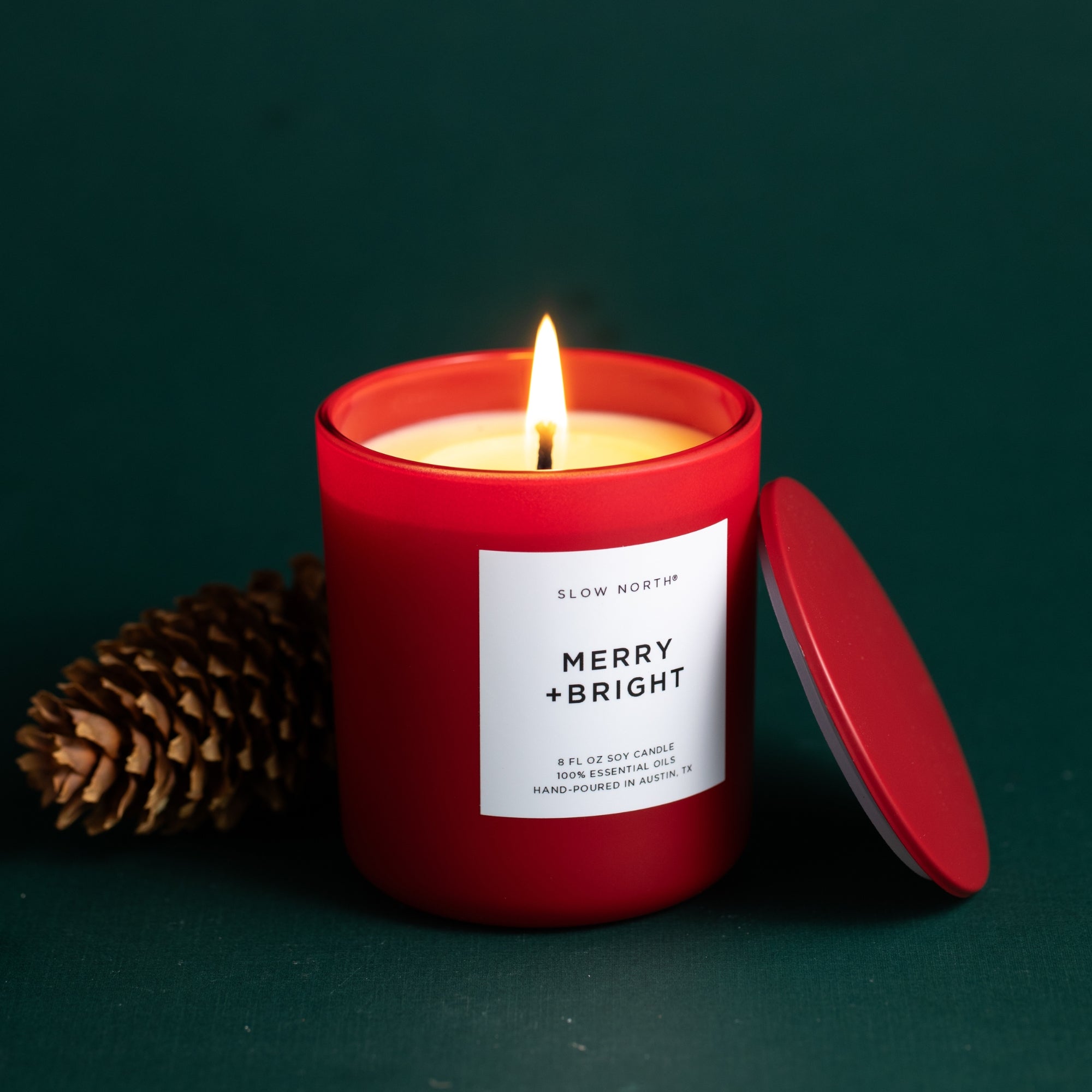 Red candle with a lit wick and 'Merry + Bright' label on a dark green background