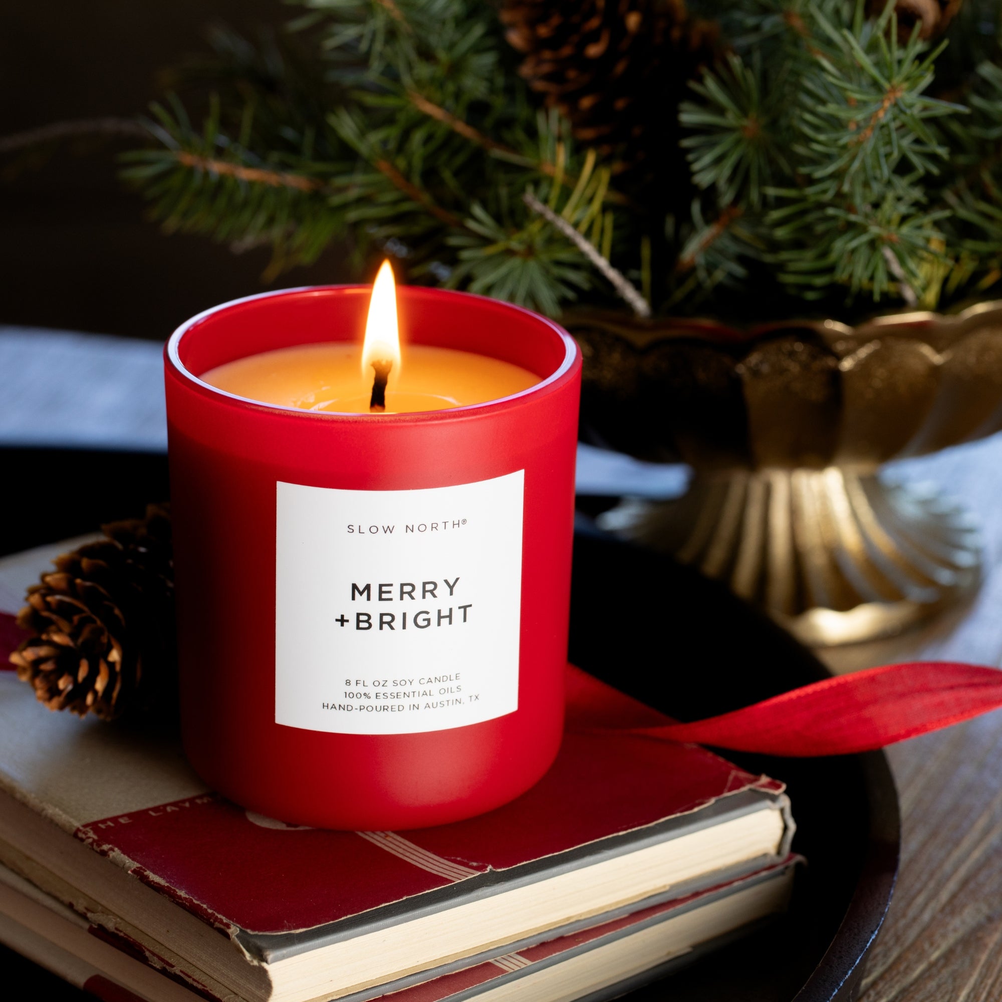 Red candle labeled 'Merry + Bright' on a stack of books with a Christmas tree in the background