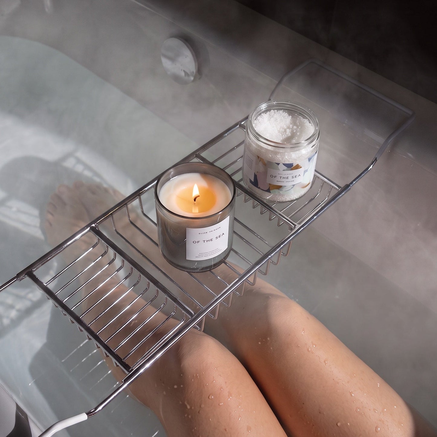 A metal bath caddy holding a candle and bath salts sits on a bathtub with a person's legs visible in the water. The scene is dimly lit with steam or mist around the tub, creating a relaxing spa-like atmosphere.