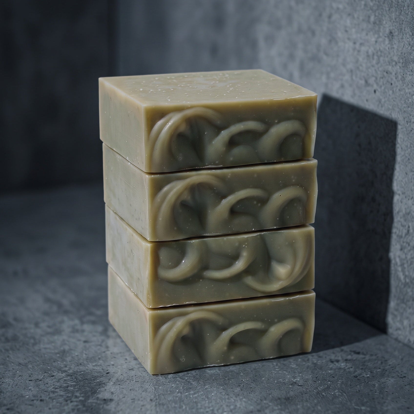 Four stacked bars of handmade soap in a warm beige color, featuring a raised circular interlocking pattern on the top surface, set against a dark textured gray background.