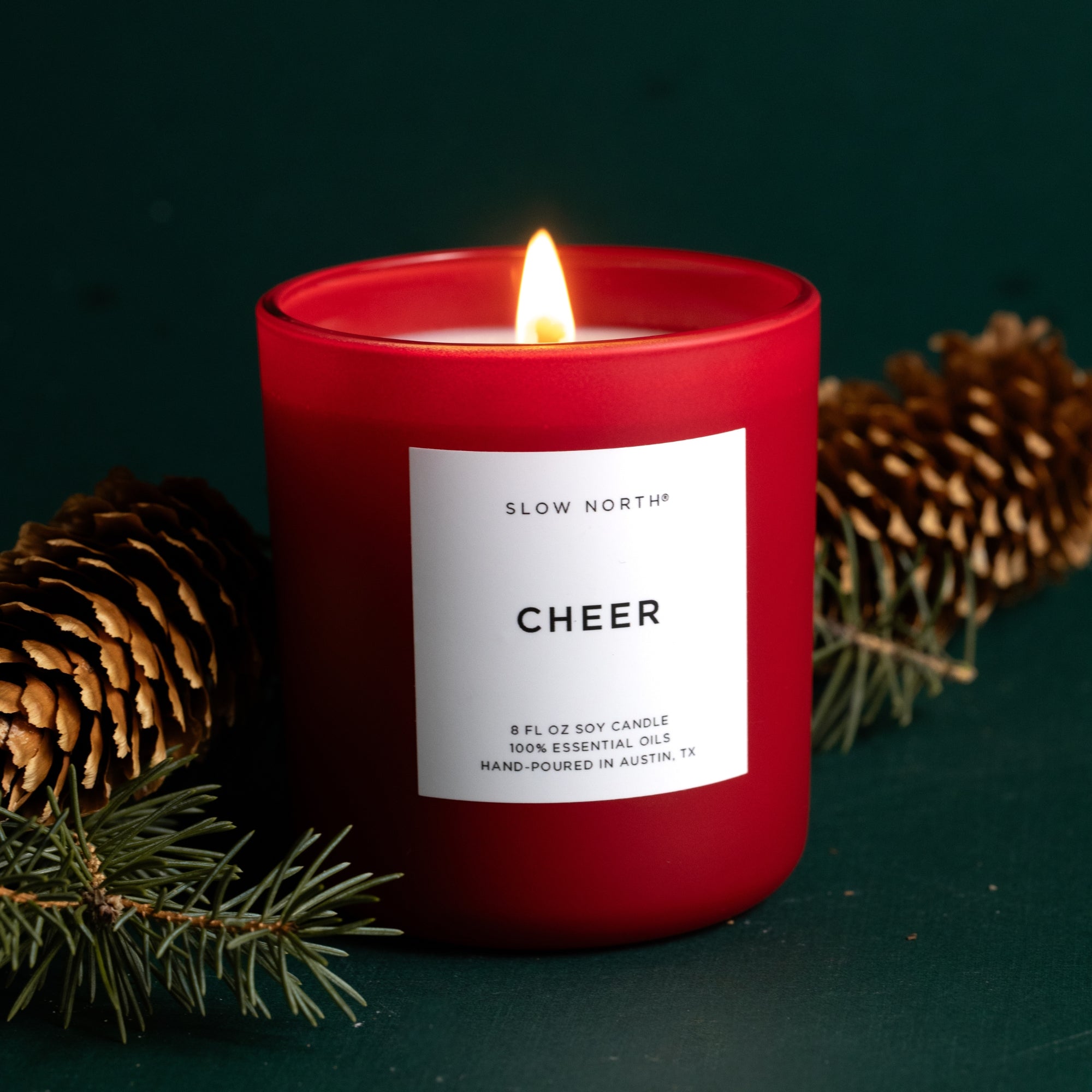 Red candle labeled 'Cheer' with pine cones and branches on a dark background