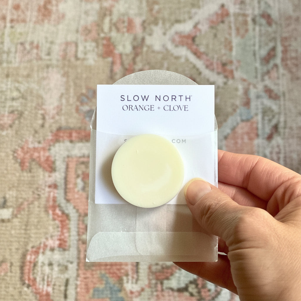 Hand holding a round wax melt scent sample labeled 'Slow North Orange + Clove' against a patterned fabric background