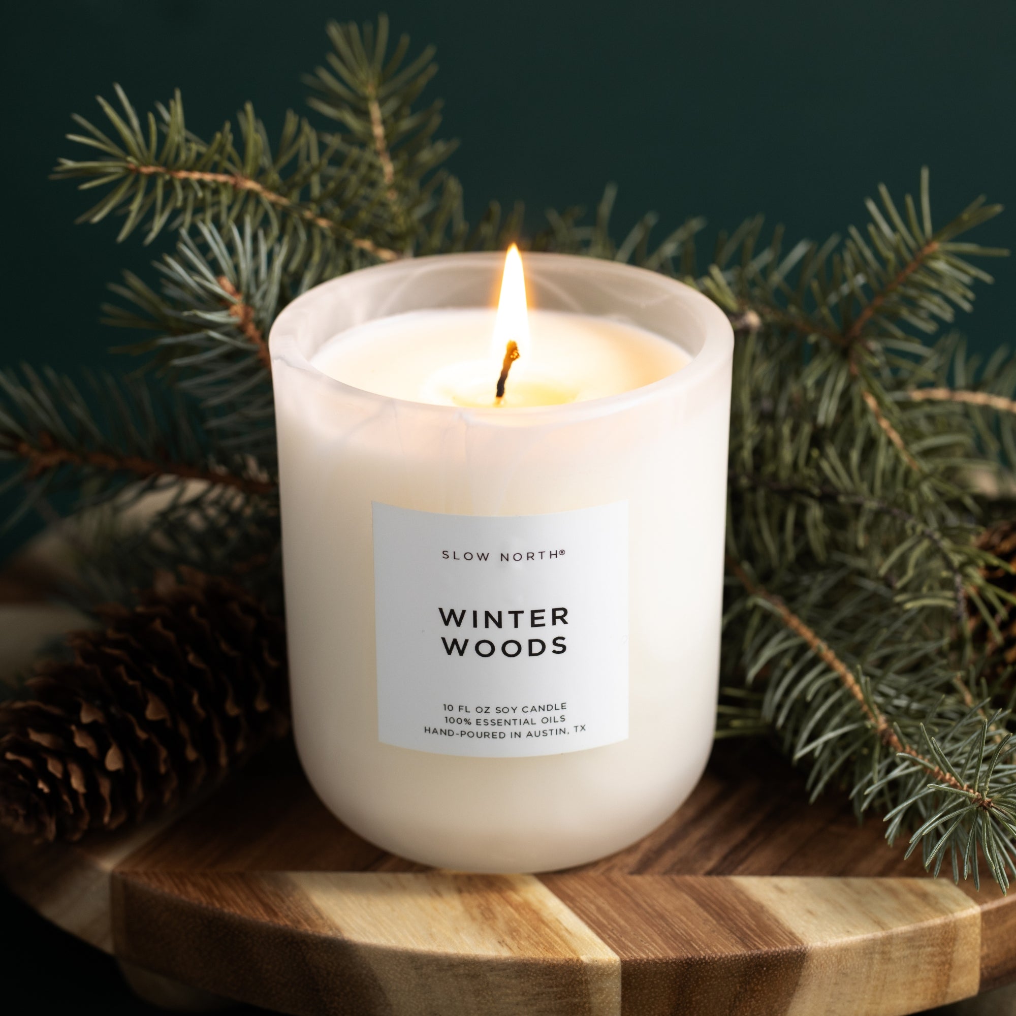 Candle labeled 'Winter Woods' with pine branches on a wooden surface