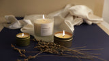 Soy wax candles lit together. Moonglow 8 ounce, 6 ounce gold travel tin, and 2 ounce mini candle together with silk fabric in background and brown floral stems near base of candles. By Slow North