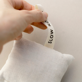 Woman's hand pinching the SLOW NORTH loop on a lavender + cedar off-white sachet