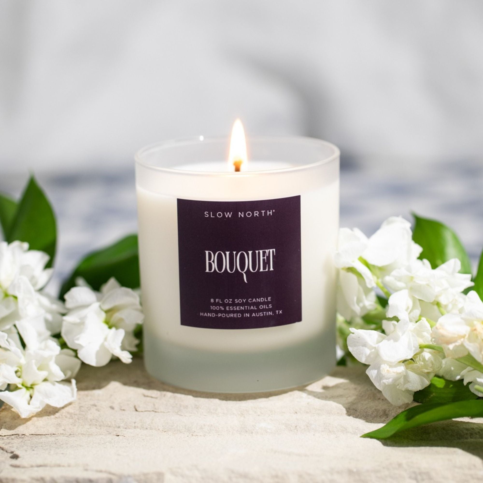 Candle with a 'Bouquet' label surrounded by white flowers on a light surface