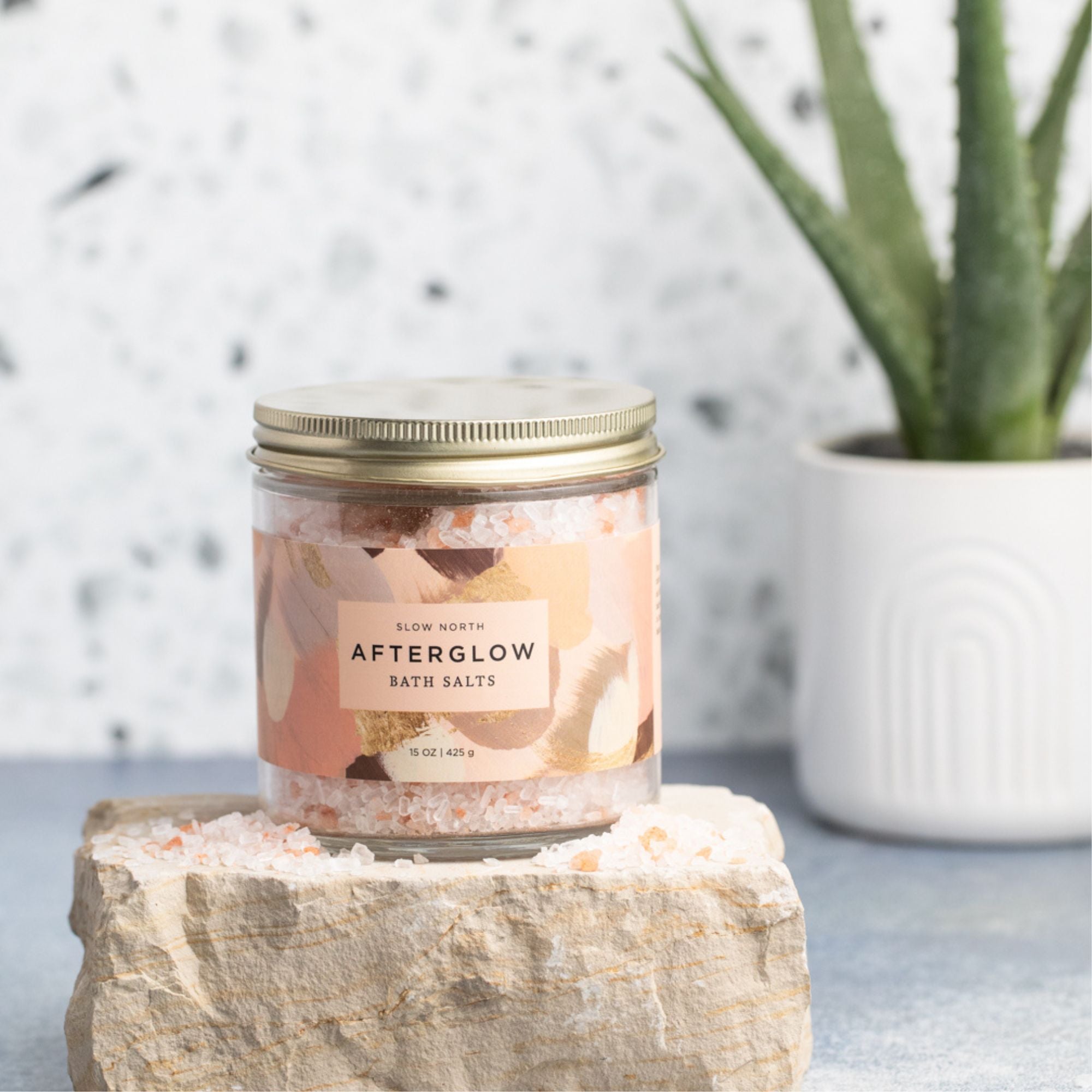 Jar of 'Afterglow' bath salts on a stone surface with a plant in the background