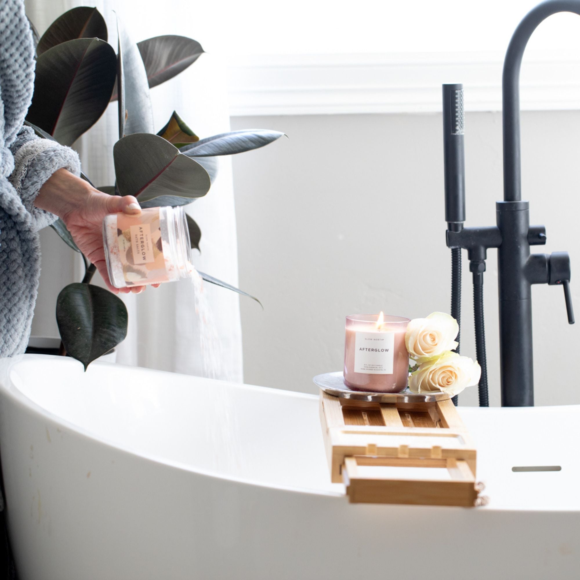 Person holding a bottle of bath product next to a lit candle and flowers in a bathtub.