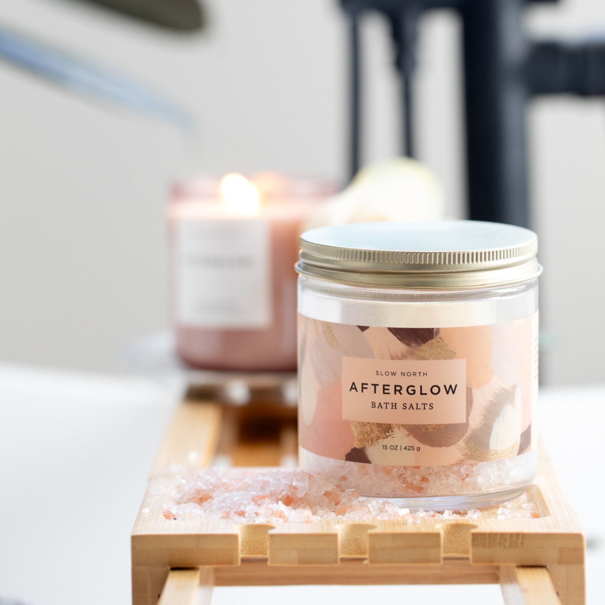 Jar of 'Afterglow' bath salts on a wooden stand with blurred candles in the background.