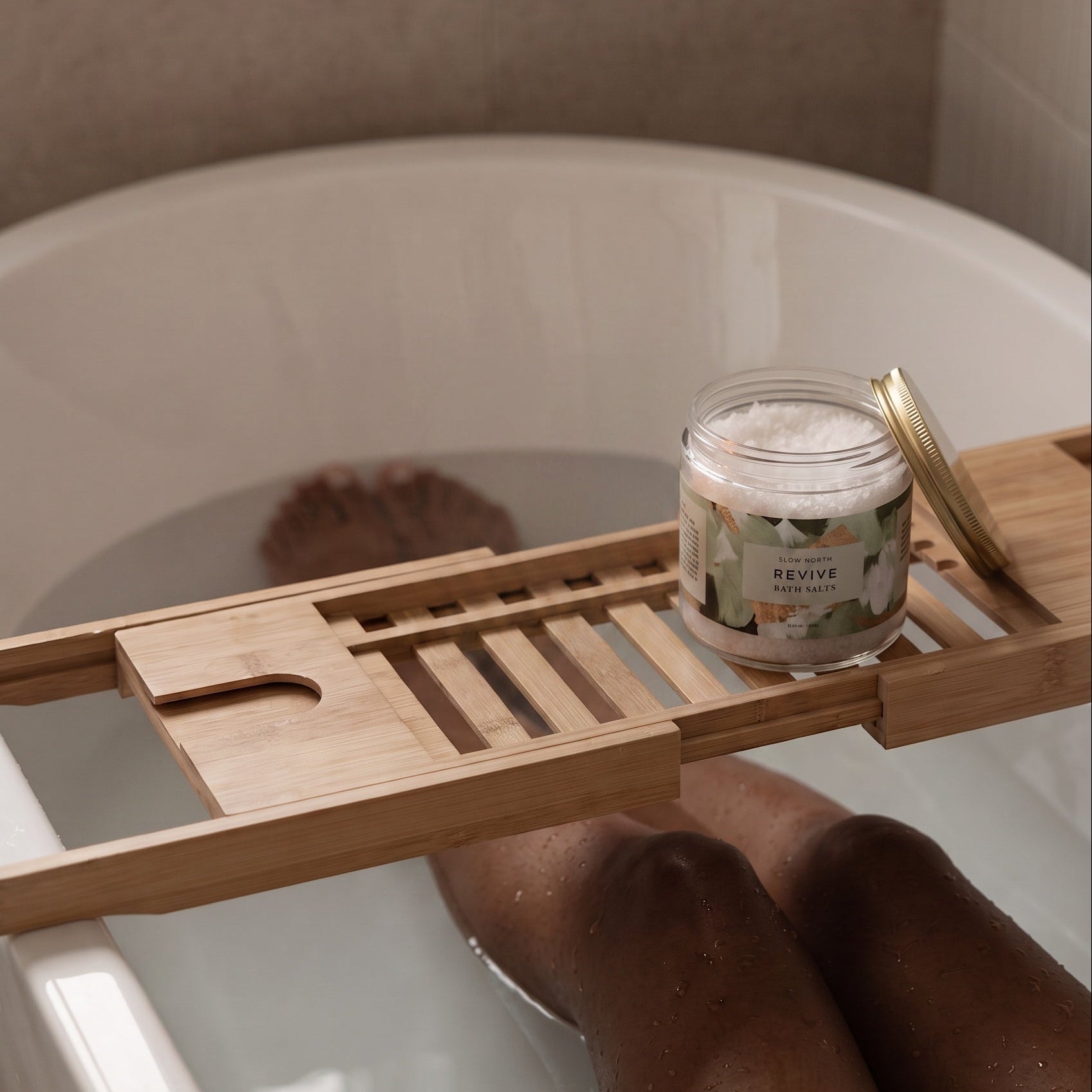 A wooden bath caddy draped over a white bathtub, holding a small jar of bath salts with a gold lid. A person's legs are visible in the water, and the background shows a tiled bathroom wall.