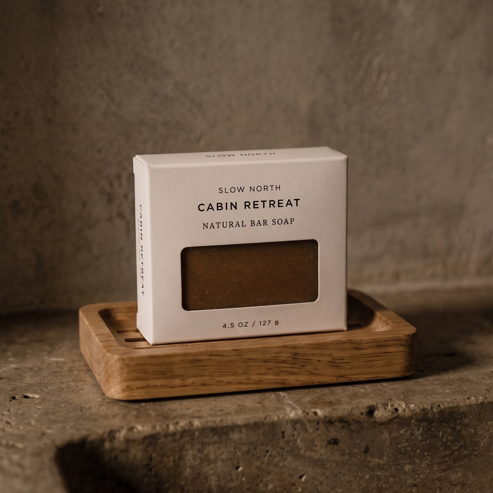 A boxed natural bar soap from Slow North Cabin Retreat sits on a small wooden tray against a textured concrete wall and floor.