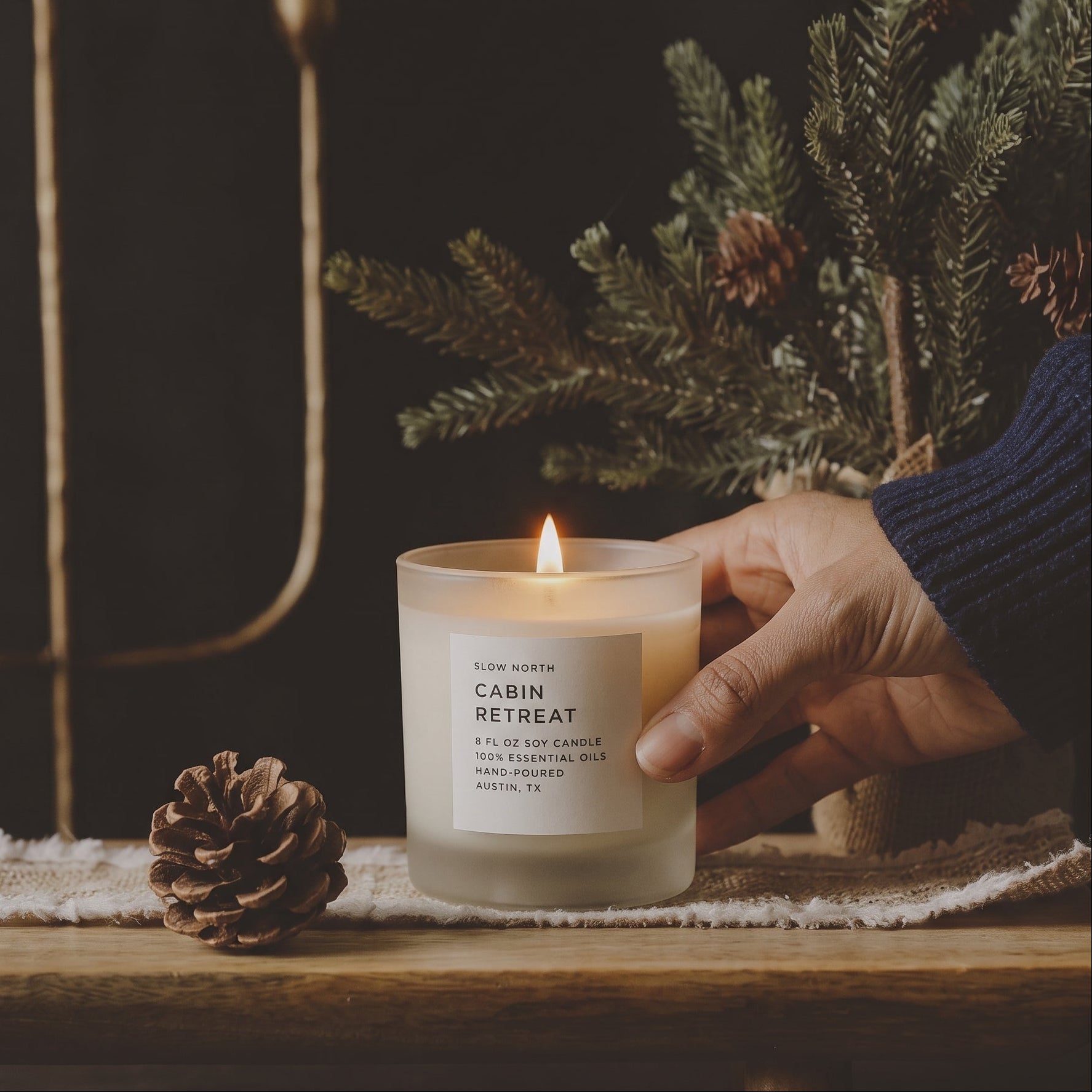 A lit candle in a frosted glass jar sits on a wooden surface, held by a hand wearing a navy sweater. The label reads 'CABIN RETREAT' 