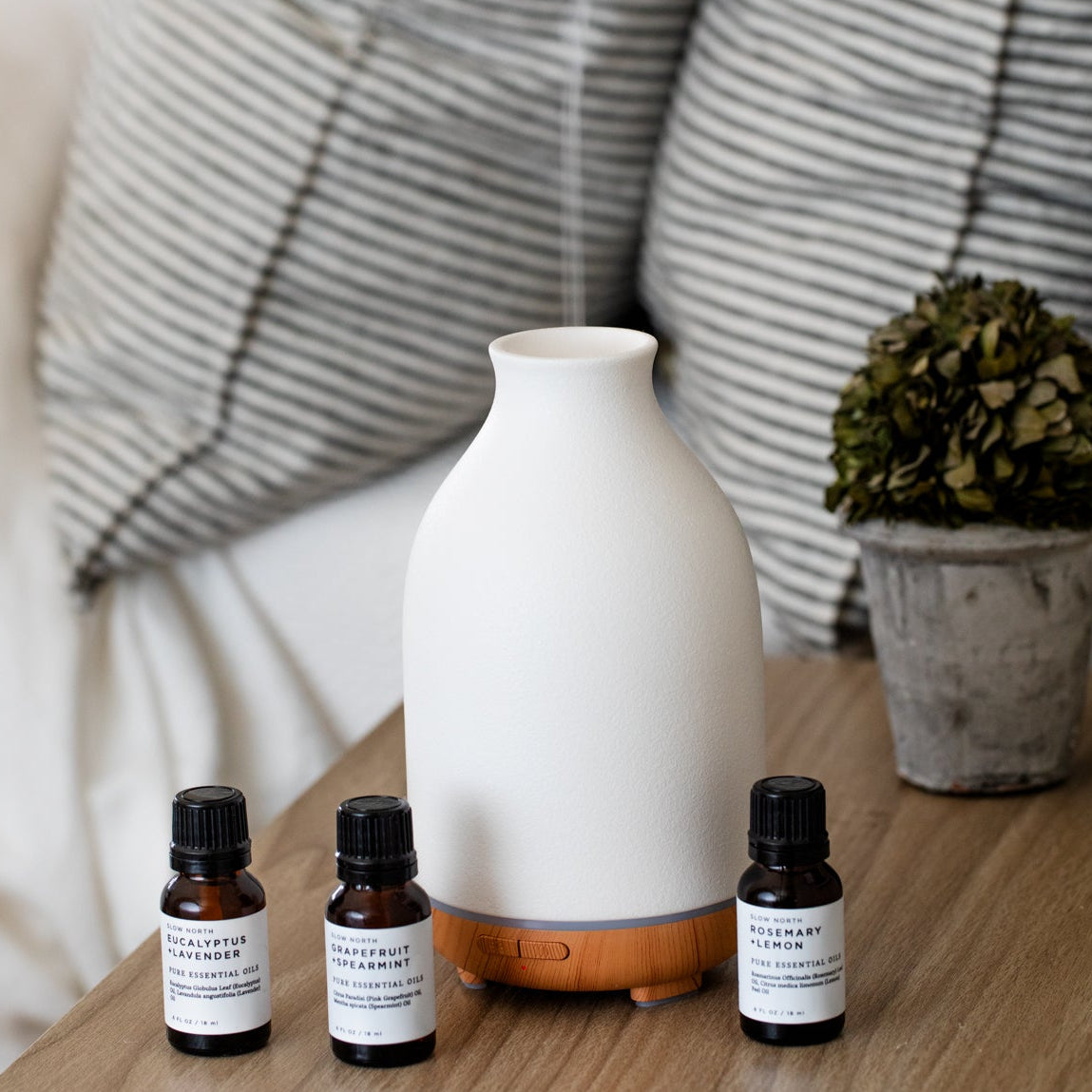 White diffuser and essential oil bottles on a wooden surface with striped cushions in the background