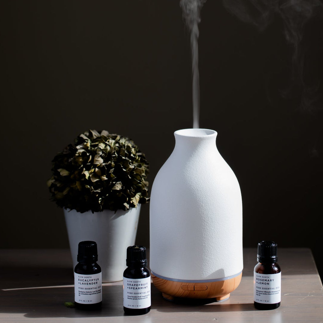 White diffuser with steam, decorative pot, and essential oil bottles on a dark background