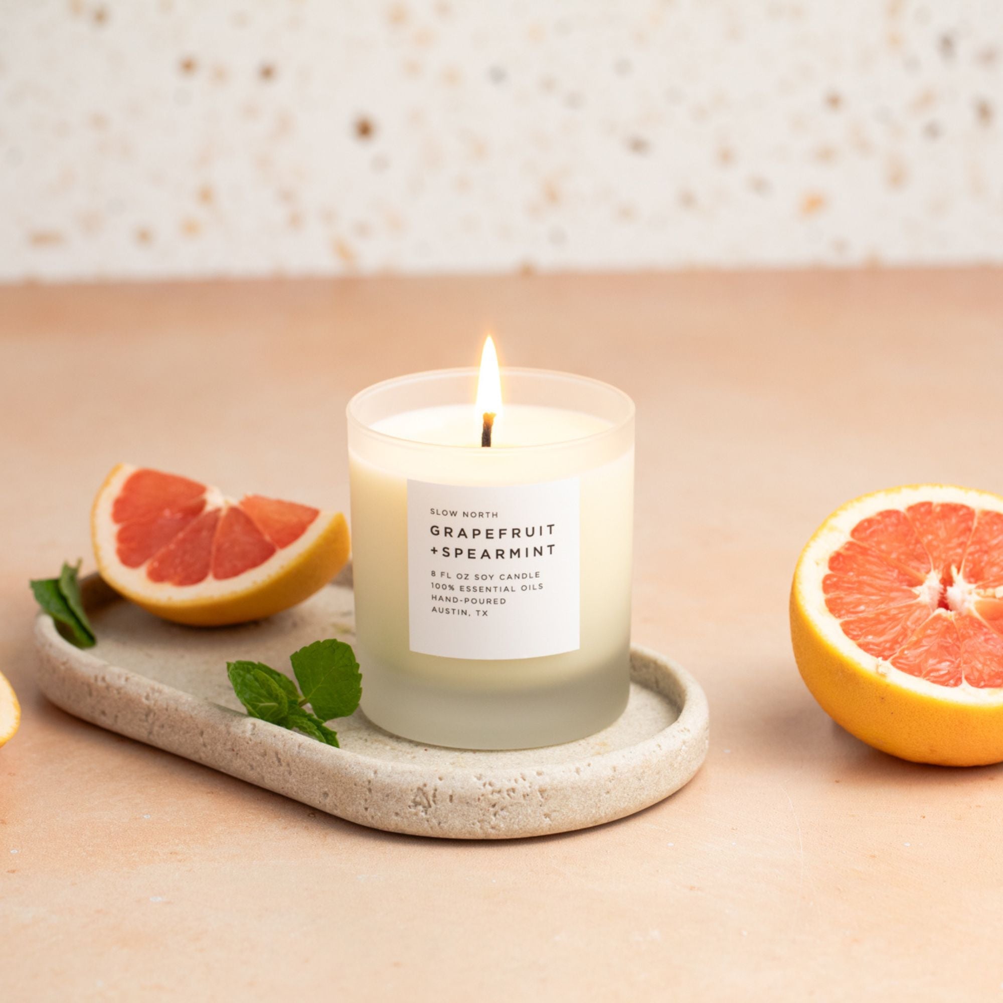 Candle labeled 'Grapefruit + Spearmint' on a stone tray with grapefruit halves and leaves on a wooden surface.