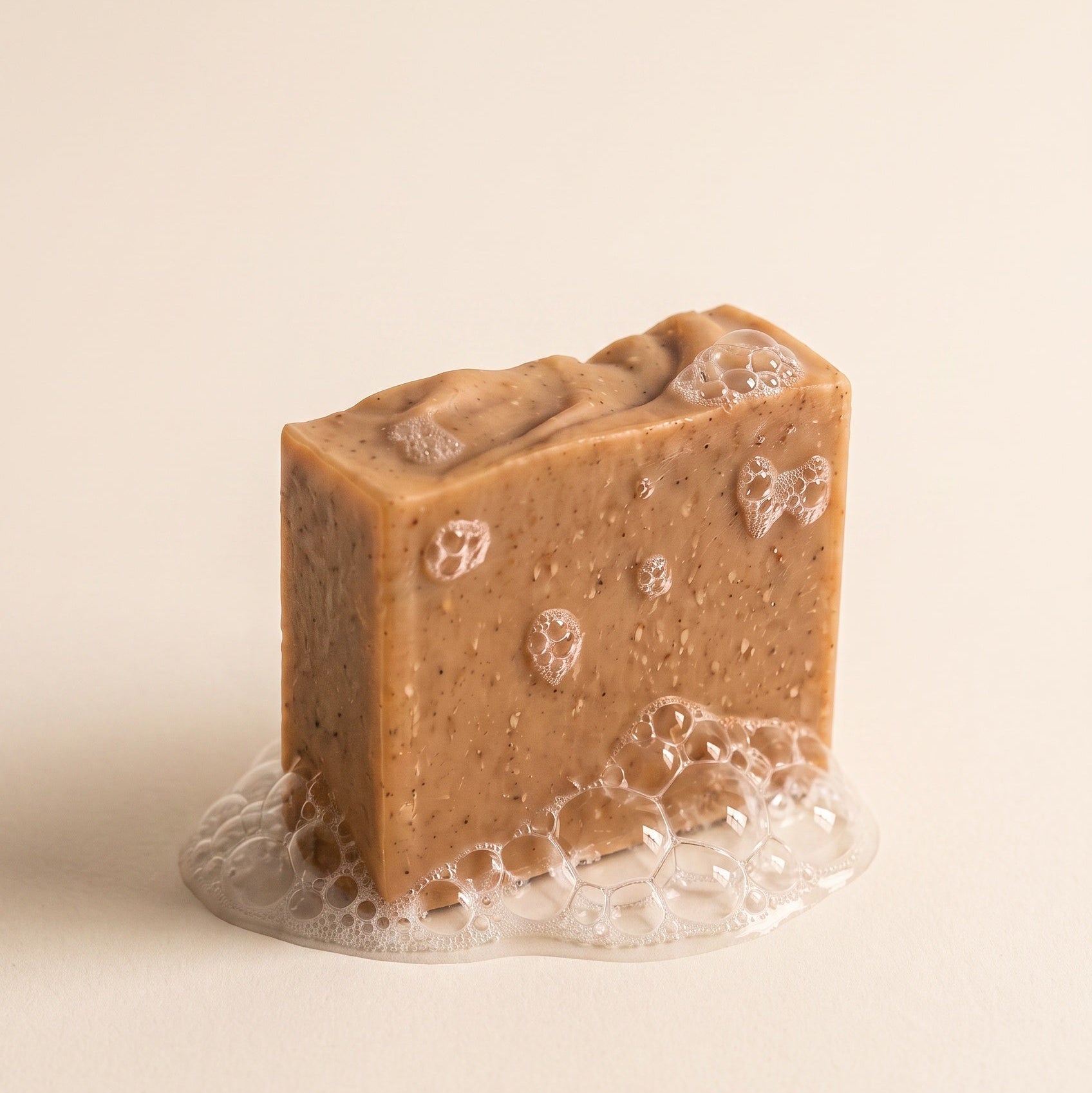 A rectangular bar of brown soap with a textured, speckled surface and small embedded bubbles, standing upright on a bed of clear foam against a warm cream background.