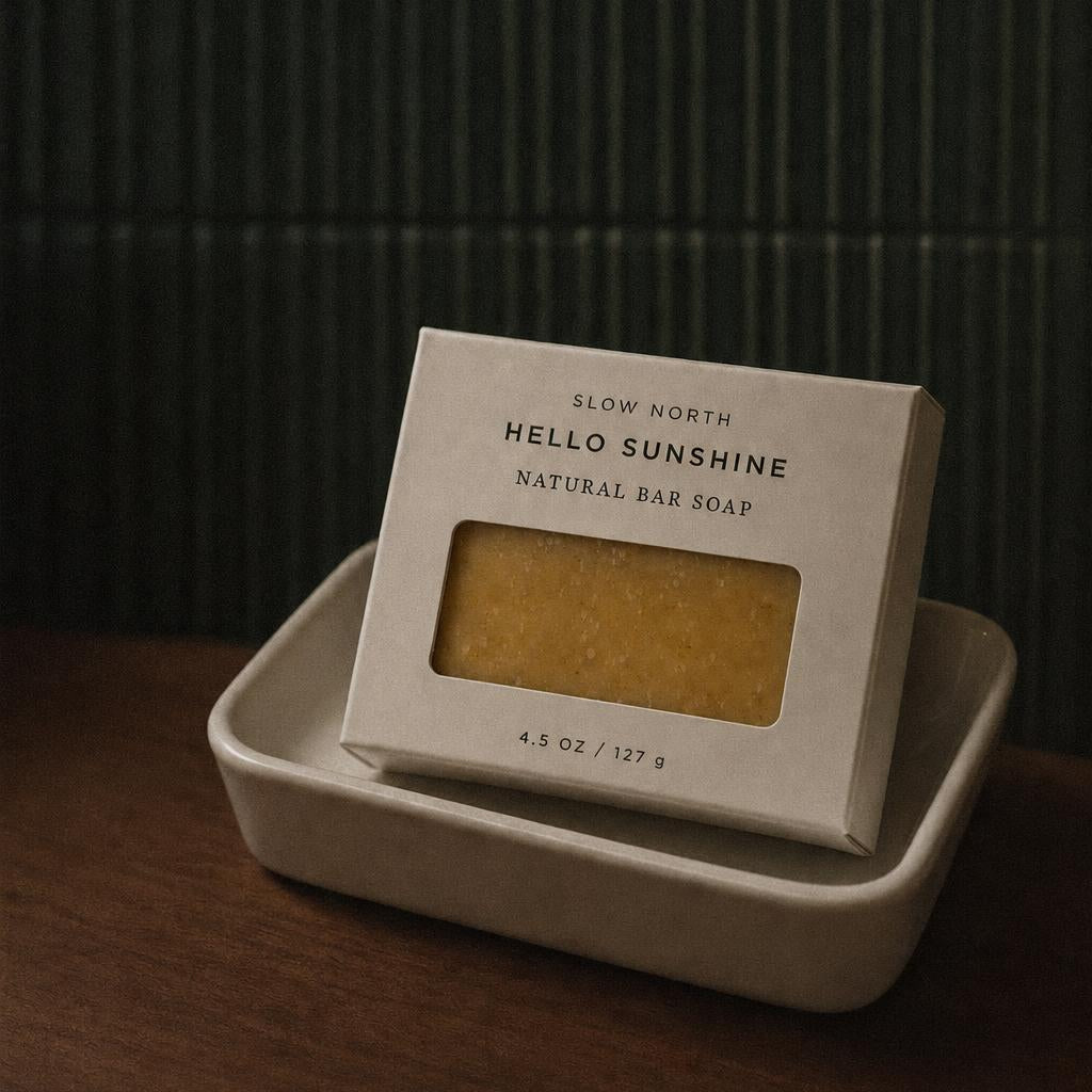 A boxed natural bar soap labeled 'Hello Sunshine' by Slow North, resting in a small beige dish on a wooden surface against a dark green paneled background.