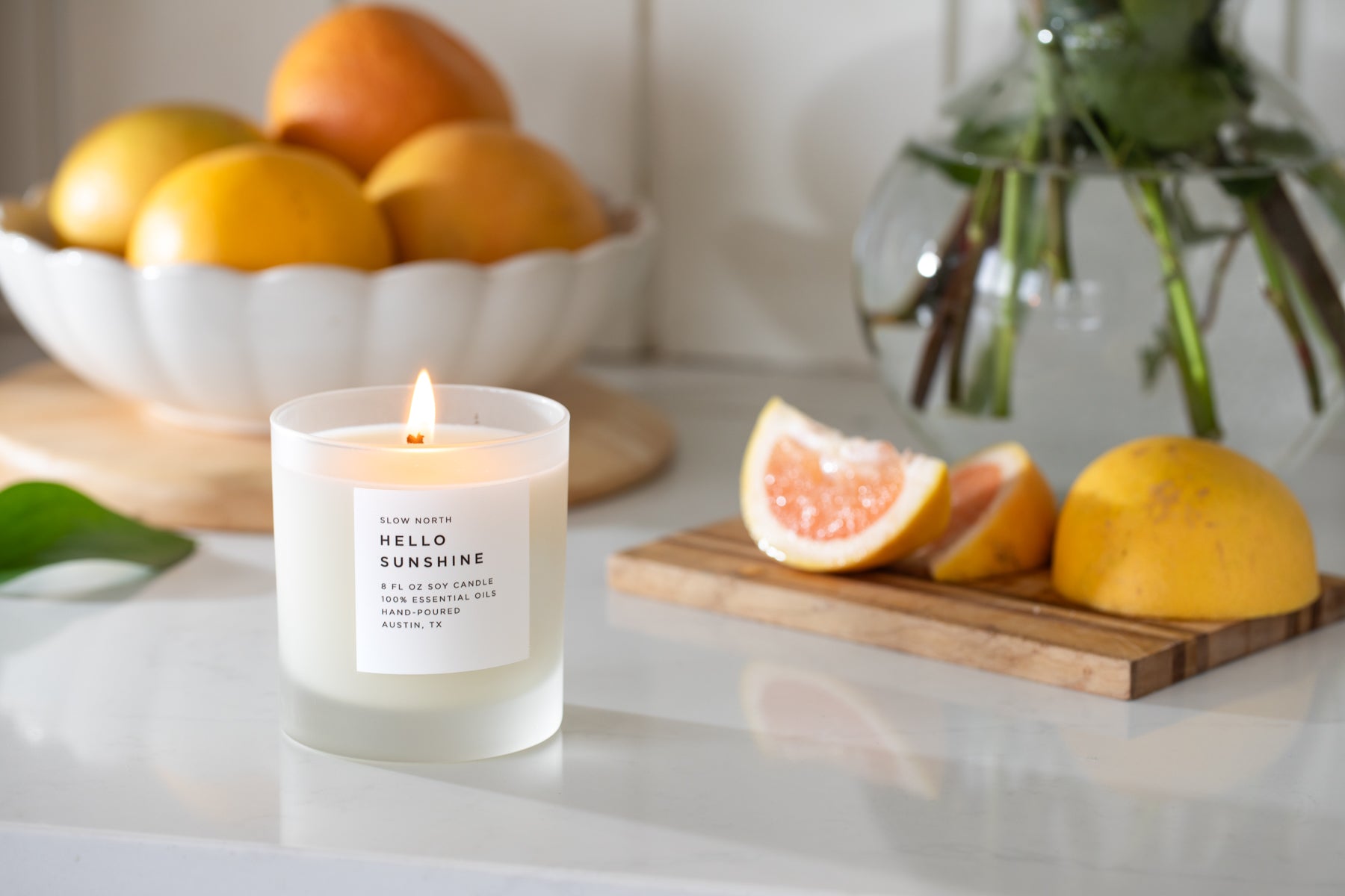Candle labeled 'Hello Sunshine' on a surface with fruits and plants in the background