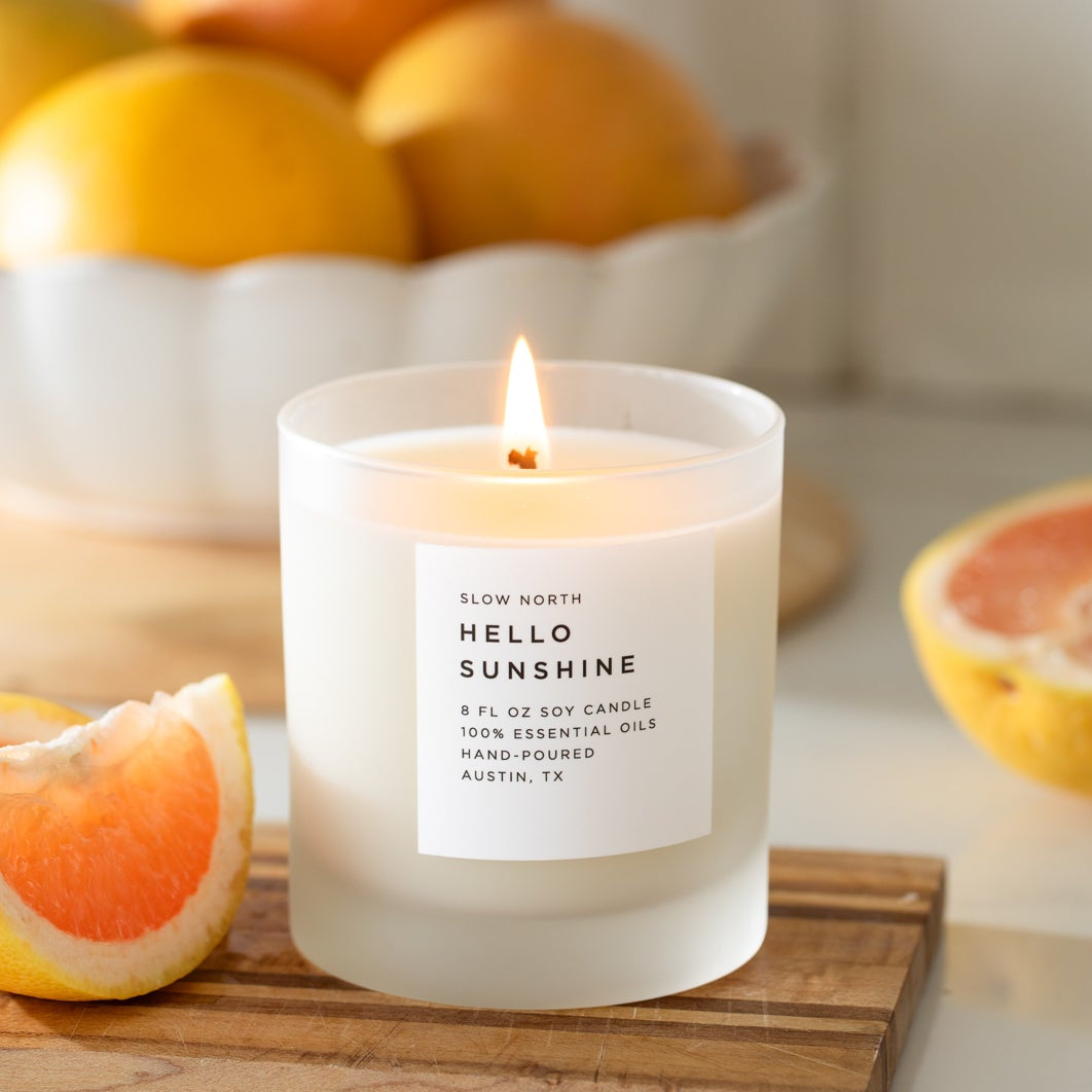Candle labeled 'Hello Sunshine' on a table with fruits in the background