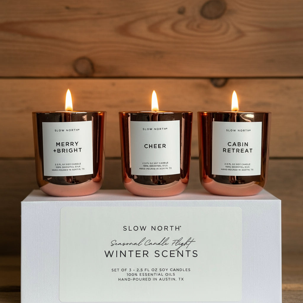 Three candles with labels 'Merry + Bright', 'Cheer', and 'Cabin Retreat' on a white box with 'Slow North' branding.