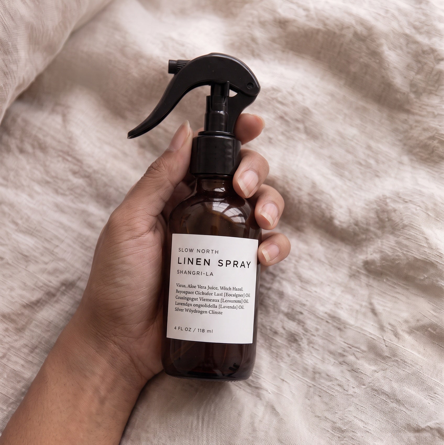 A hand holding a brown glass spray bottle of slow north linen spray with a black trigger sprayer and a white label, set against a background of textured beige linen fabric.