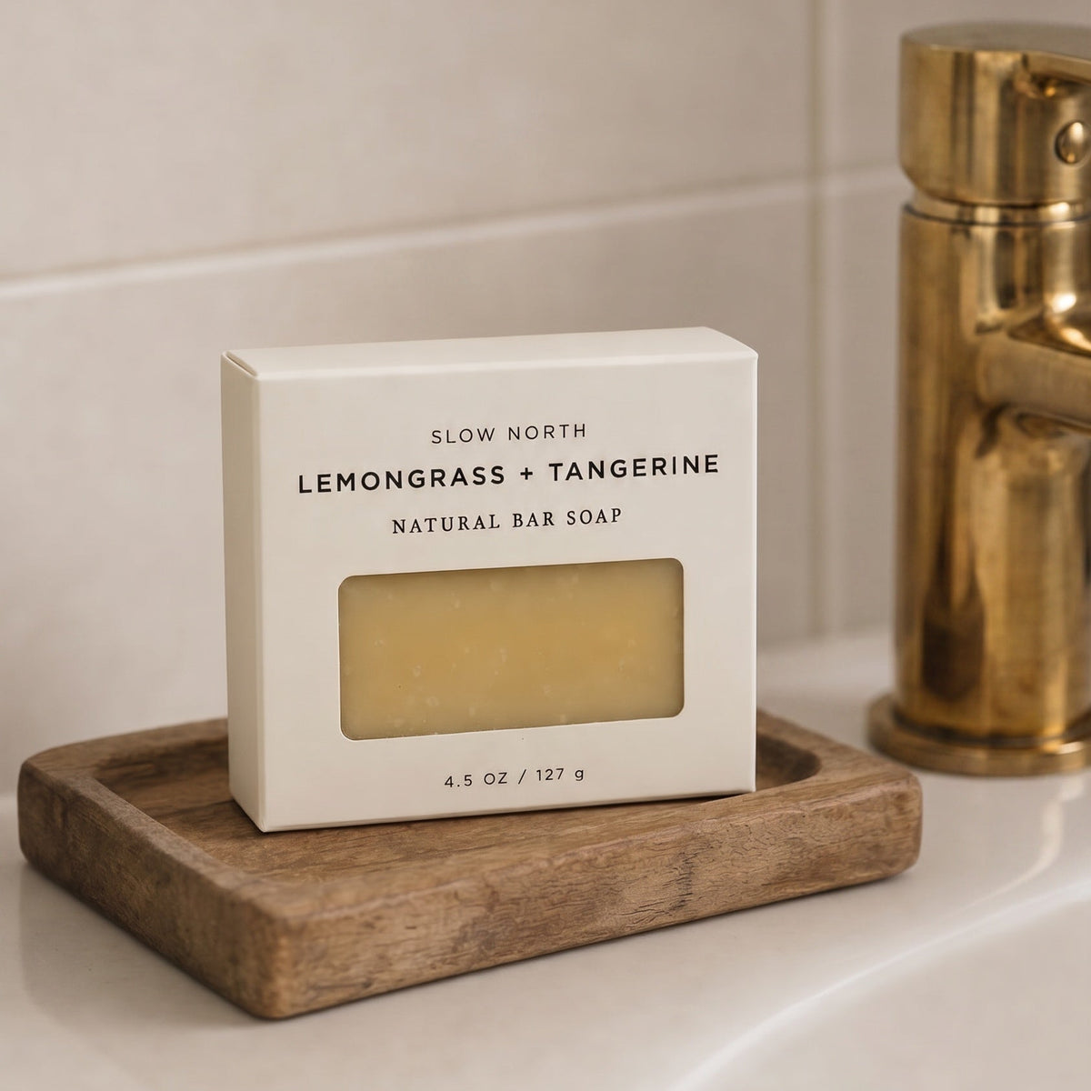 A boxed bar of soap labeled 'SLOW NORTH LEMONGRASS + TANGERINE NATURAL BAR SOAP' sits on a wooden soap dish on a bathroom counter, with a gold faucet in the background.