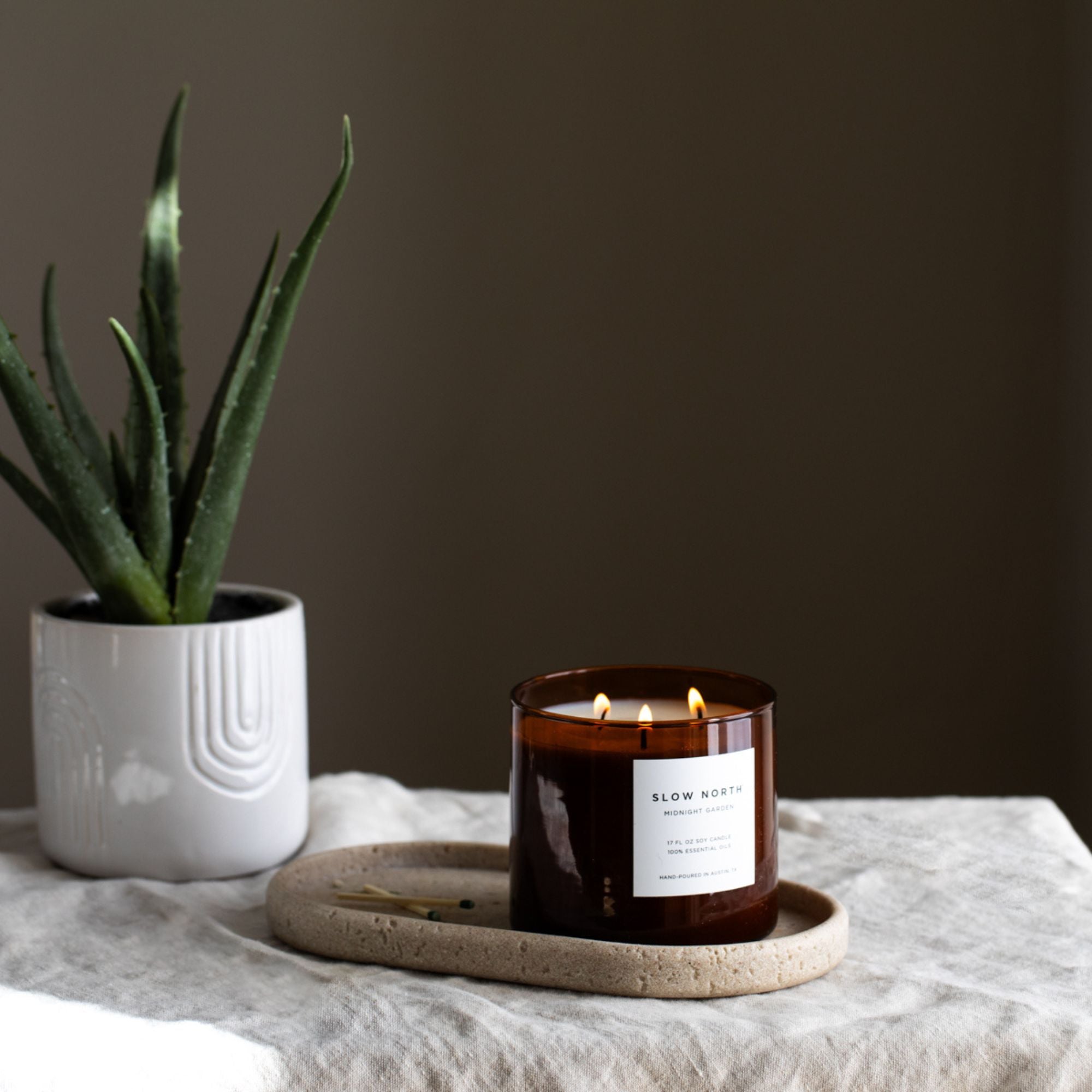 Candle with a label on a tray next to a potted plant on a table.