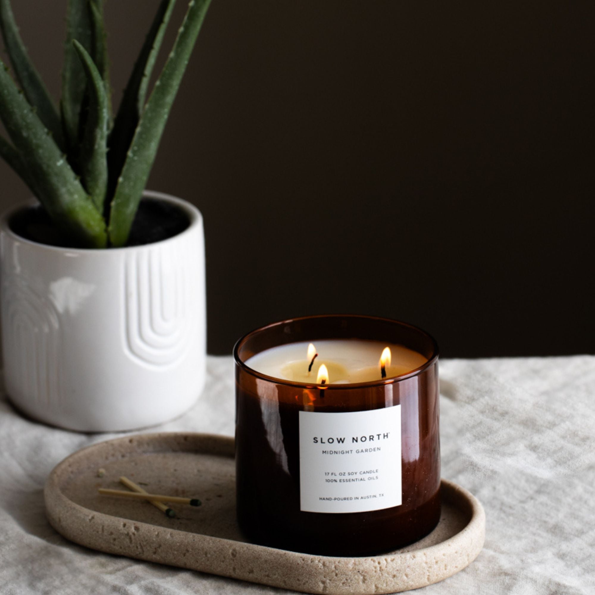 Candle in a brown jar with a label, placed on a stone coaster next to a potted plant.
