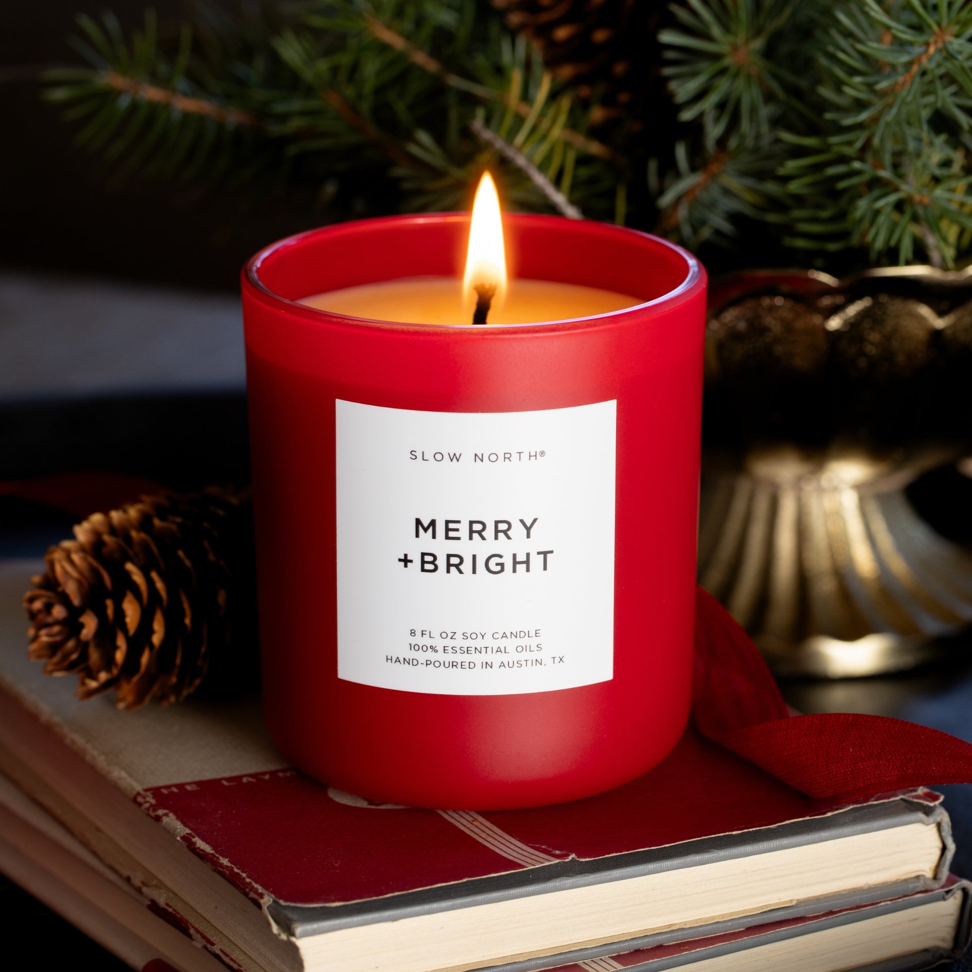 Red candle labeled 'Merry + Bright' on books with a Christmas tree in the background
