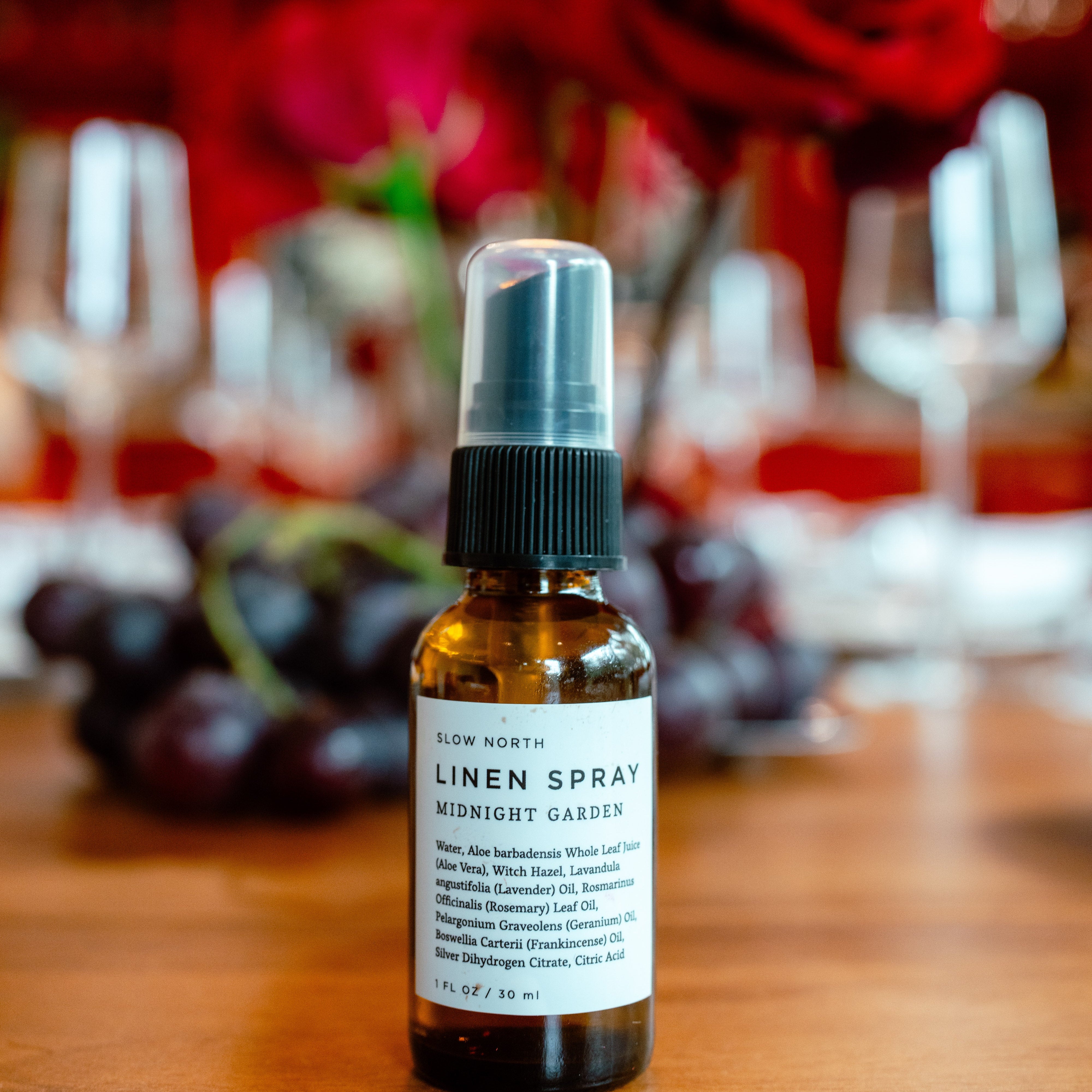 Linen spray bottle on a wooden table with blurred background of flowers and glasses.