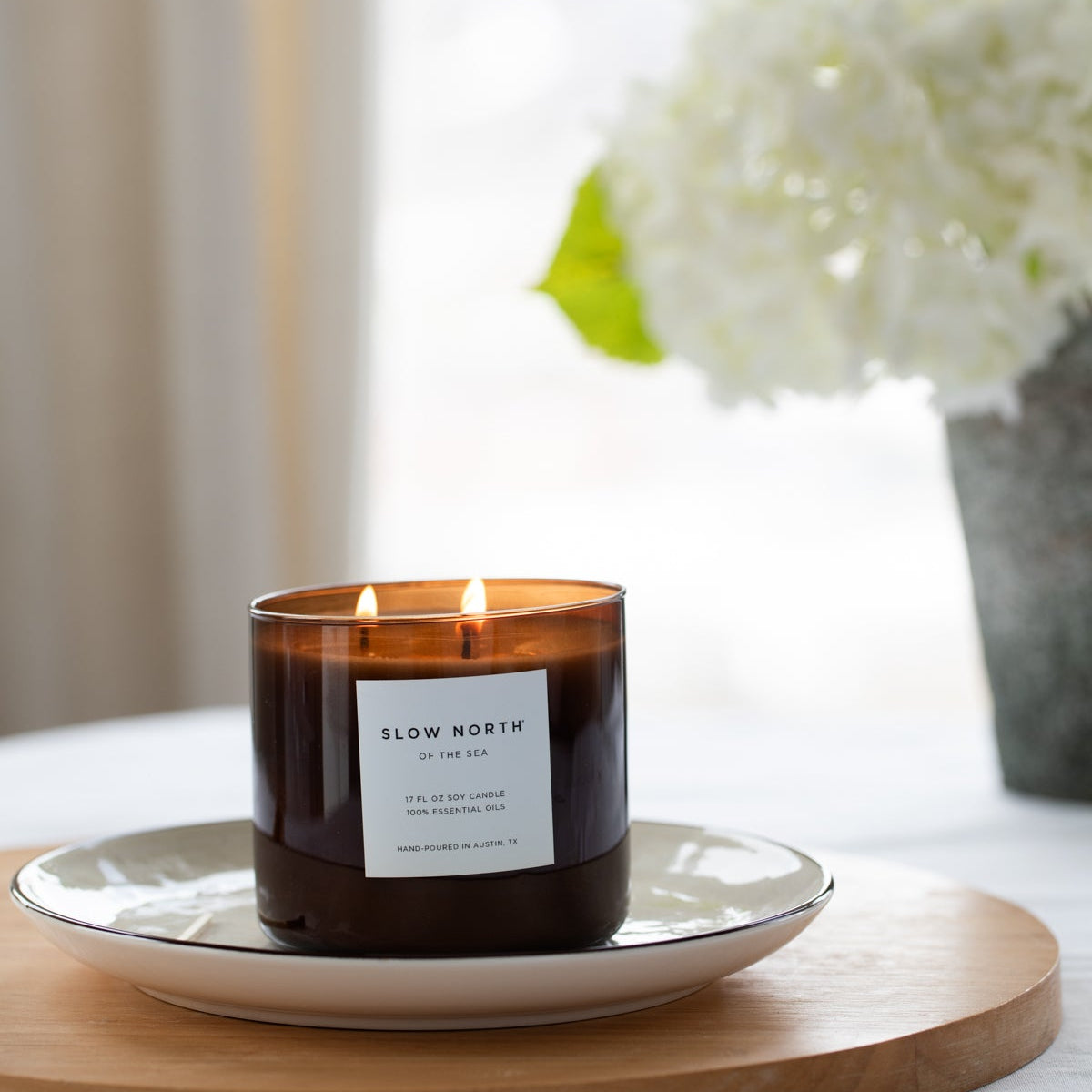 Candle with a label on a white plate on a wooden board, with white flowers in the background.