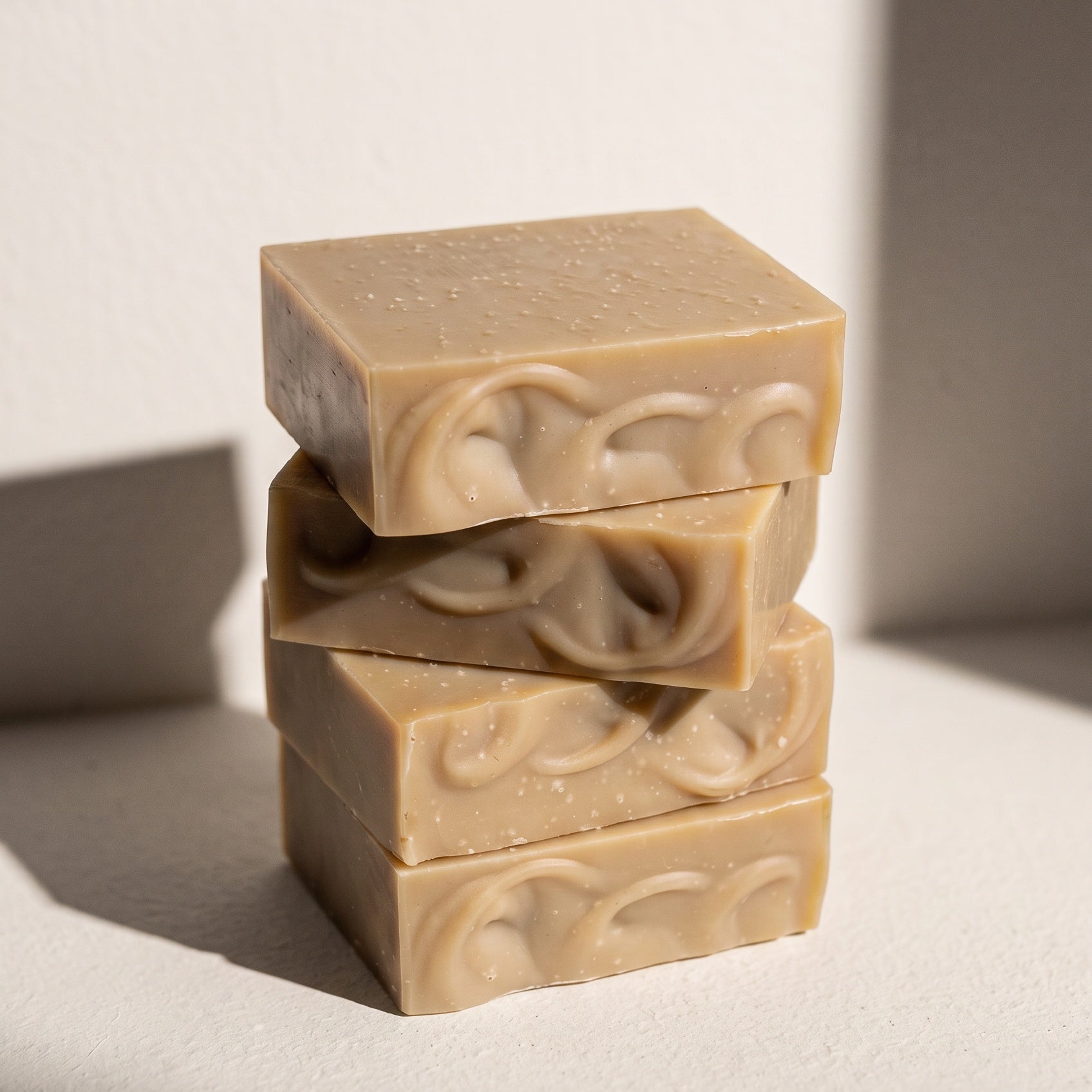 Four stacked rectangular bars of handmade soap in a warm light tan color, featuring a swirled embossed pattern on the sides, set against a minimalist off-white background with dramatic diagonal shadows.