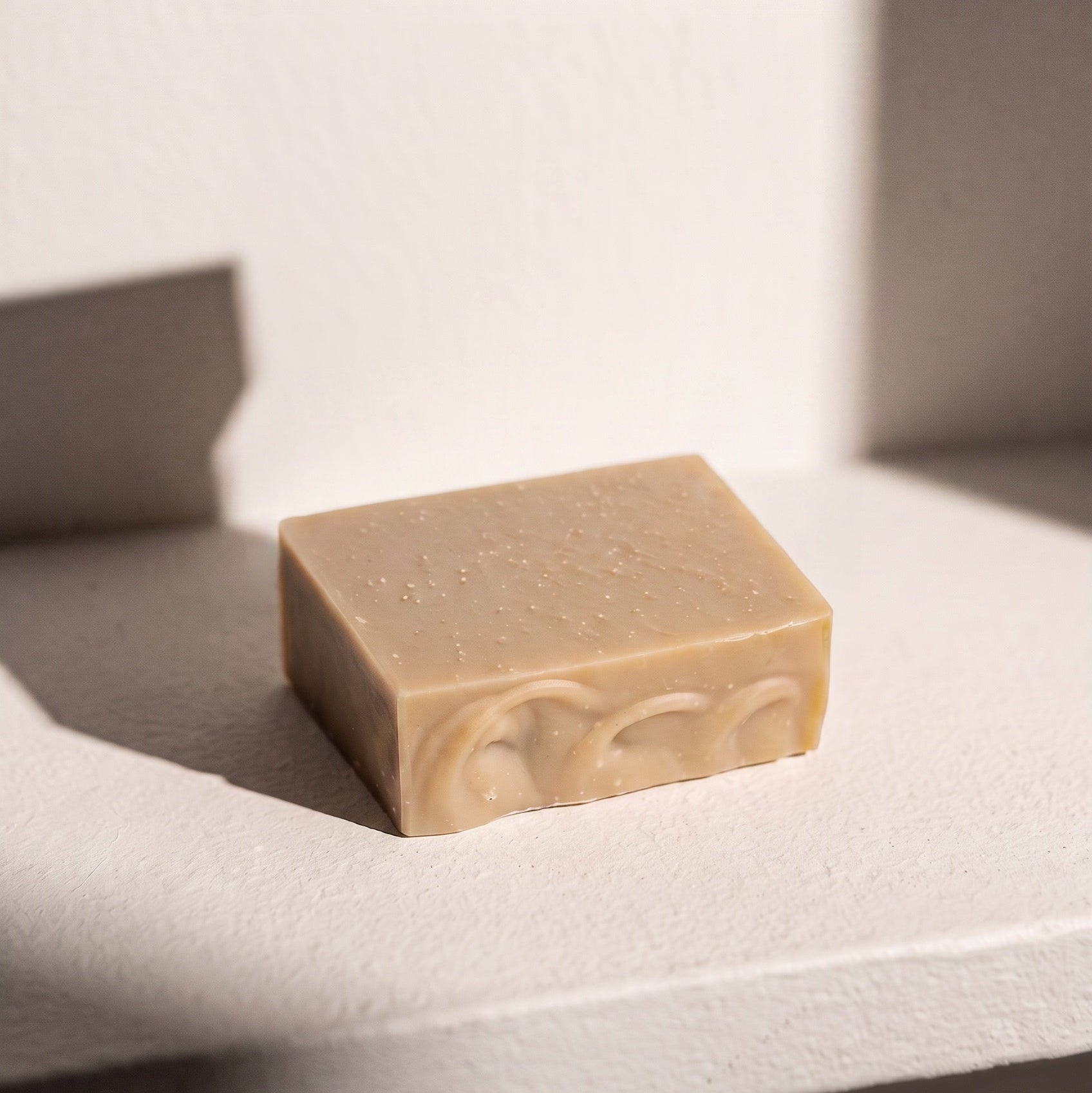 A rectangular bar of handmade soap in a light beige color with embossed swirls on one side, resting on a textured white surface under dramatic diagonal sunlight.