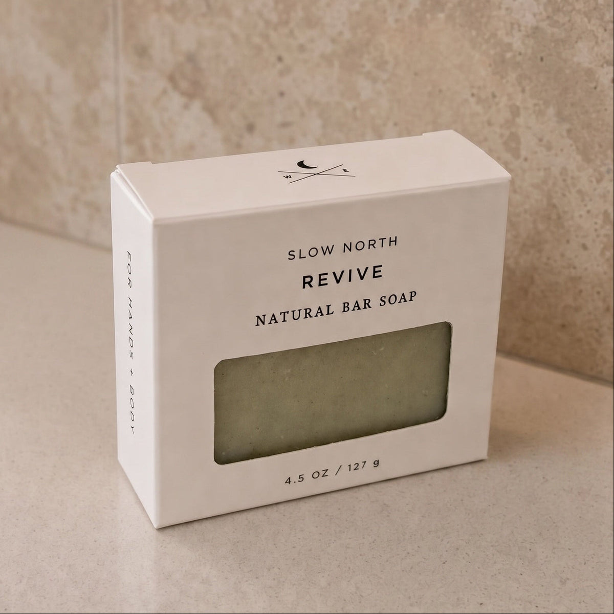 A white rectangular box of Slow North Revive Natural Bar Soap sits on a light beige stone surface against a matching stone wall background.