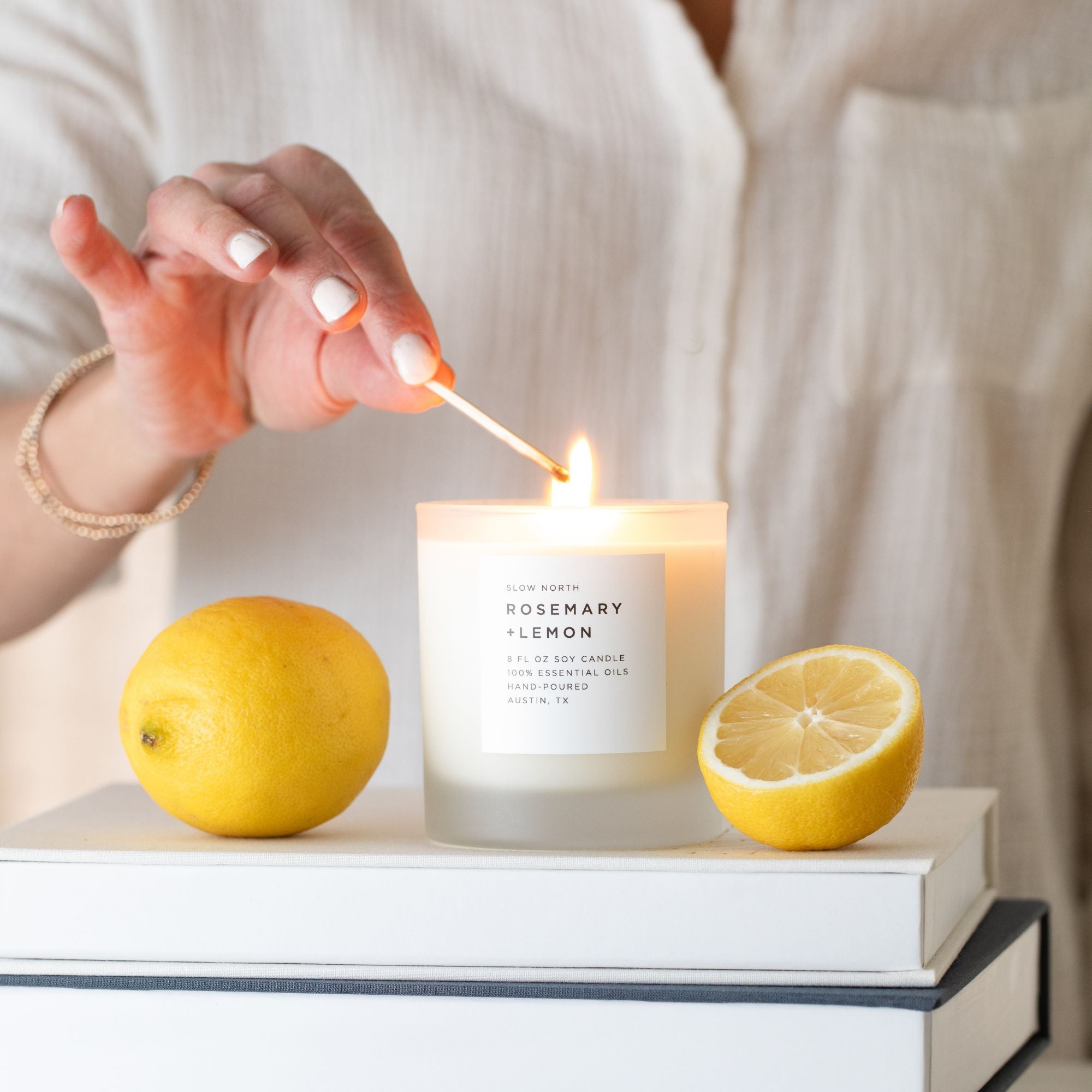 Person lighting a candle labeled 'Rosemary Lemon' with lemons nearby on a white surface.