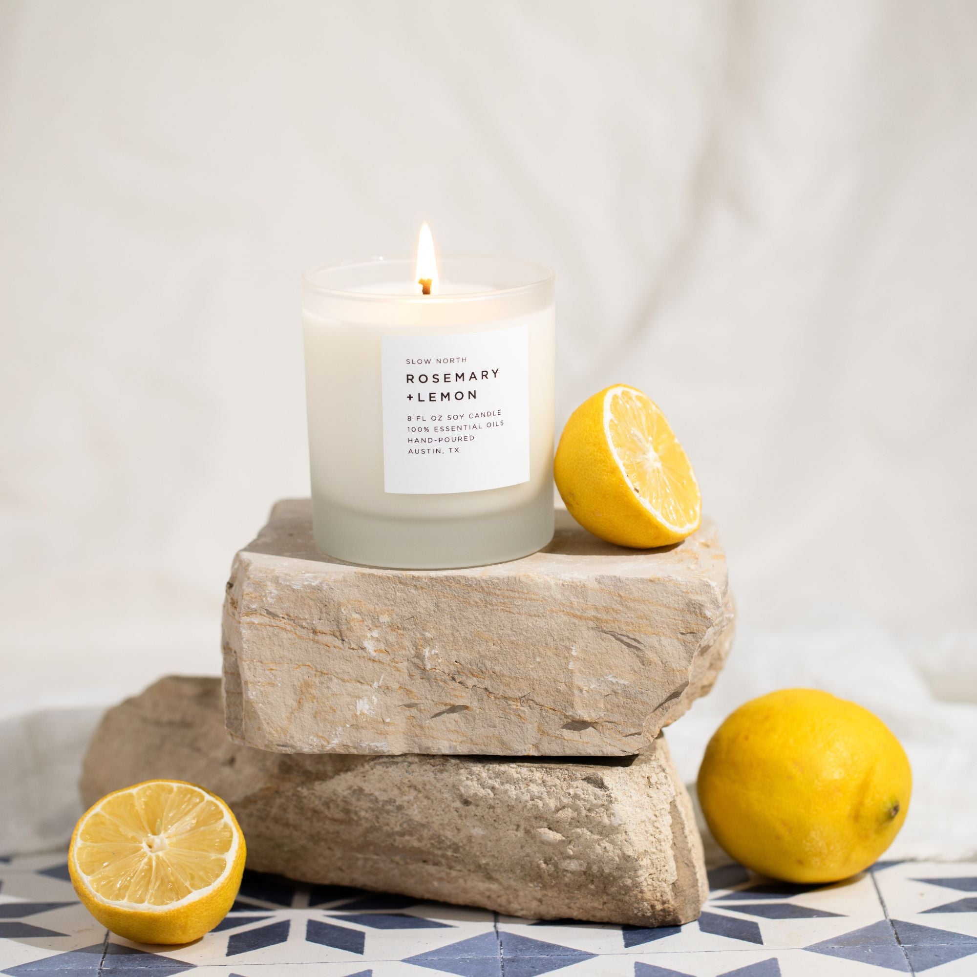 Candle labeled 'Rosemary + Lemon' on a stone block with lemons around, on a patterned surface.