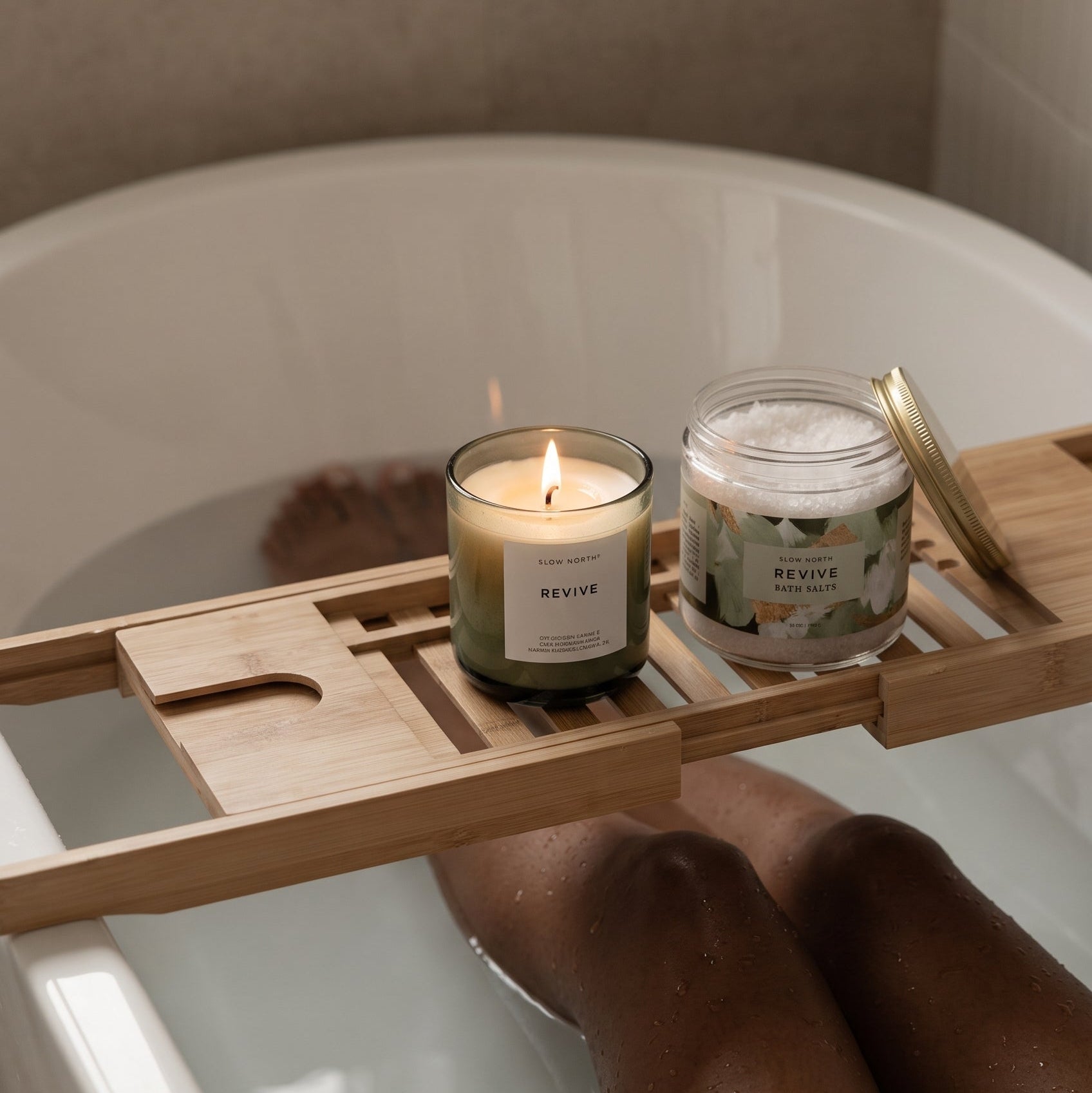 A wooden bath caddy draped over a white bathtub, holding a lit candle and a jar of bath salts. A person's legs are visible in the water, creating ripples.